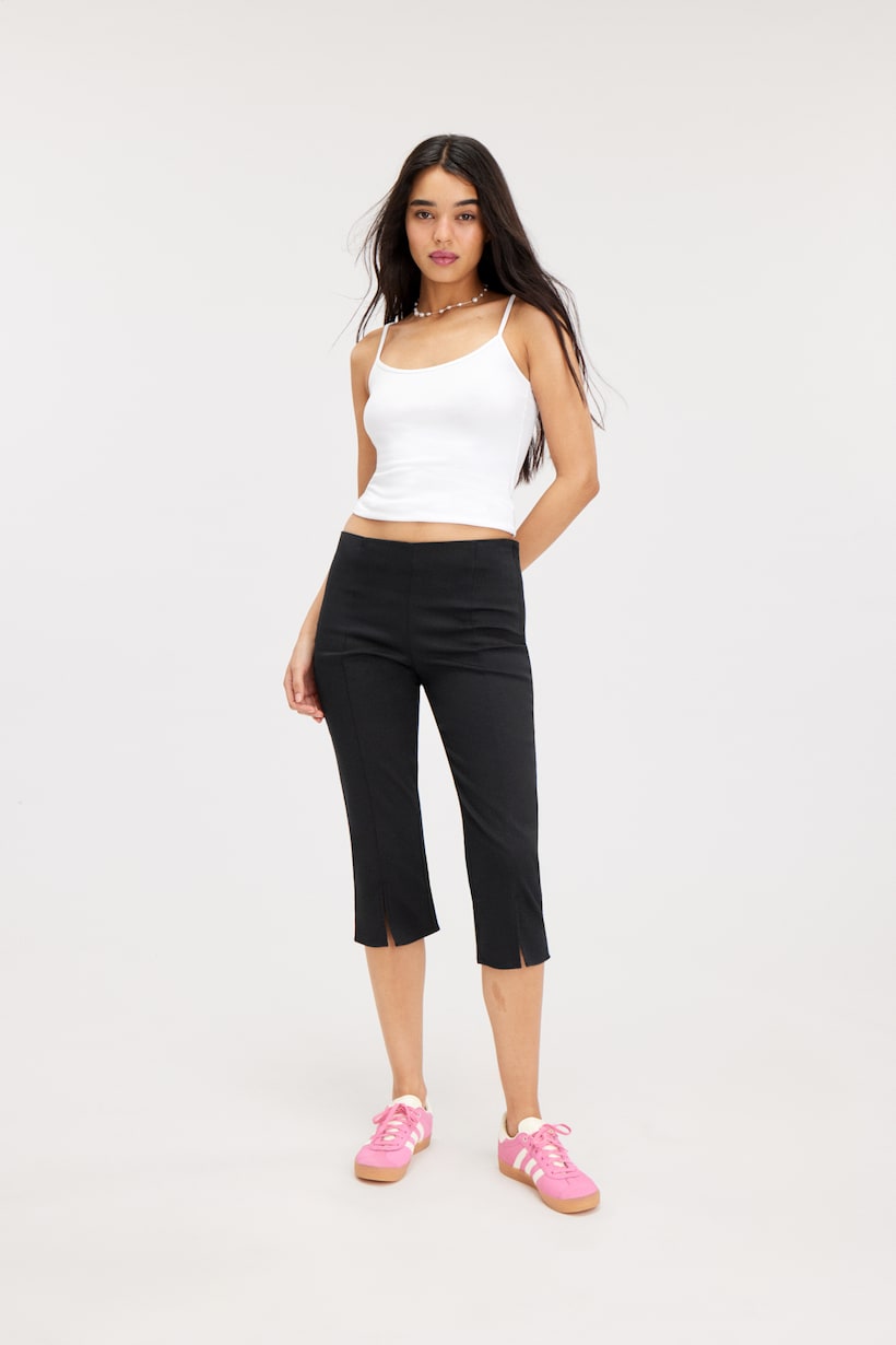 Monki black capri trousers crafted from a soft and stretchy blend of cotton, enhanced with polyester and recycled cotton. They feature a hidden side zip closure, secured with a jigger button, back darts for added structure, and front seam detailing with slitted leg openings for added style and mobility. Slim fit. Regular waist. Cropped length. Front slits.