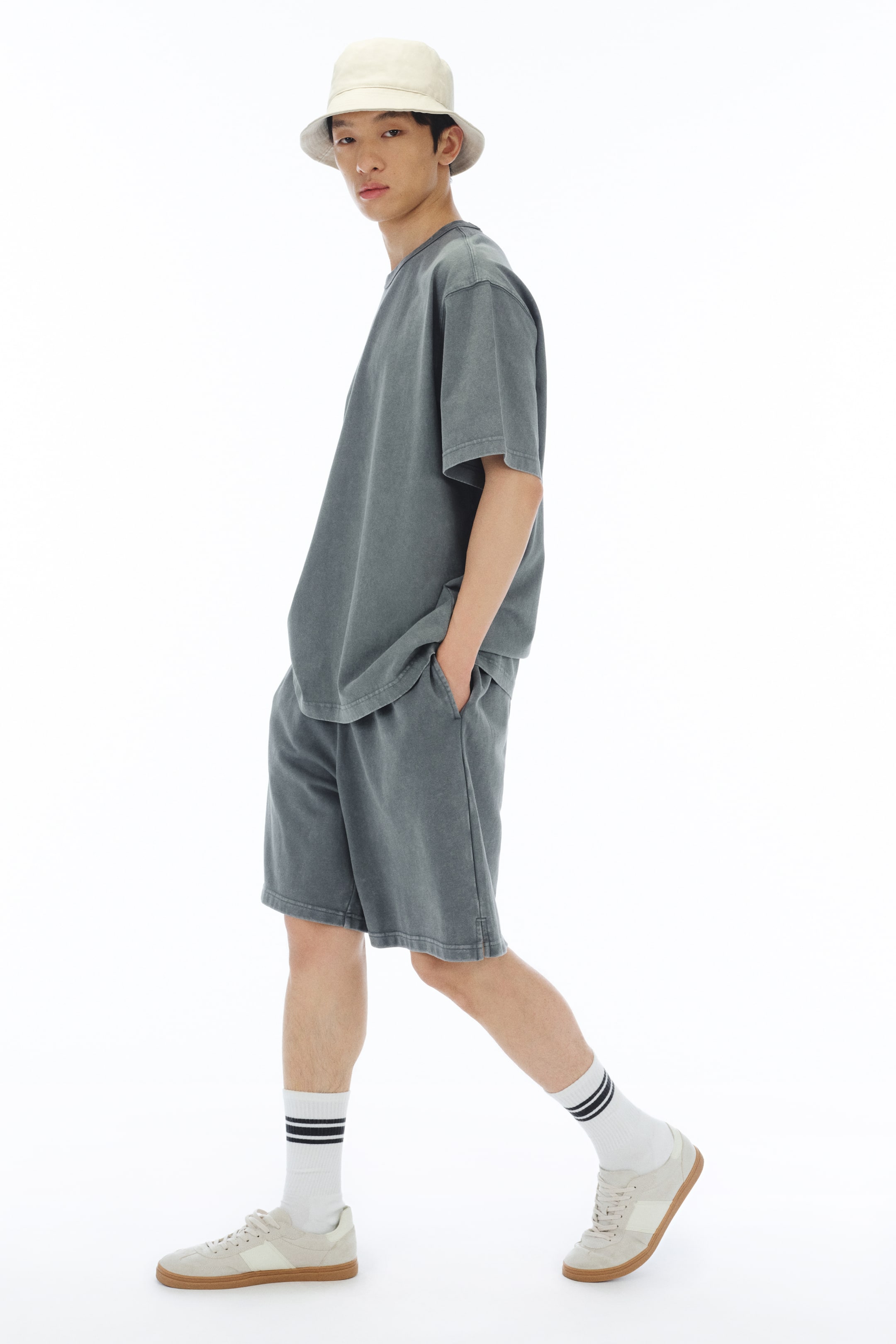 拡大画像を表示: モデルはダークグレーの半袖Tシャツとリラックスフィットのショーツのセットを着用し、ライトカラーのバケットハットをかぶっています。モデルは片手をショーツのポケットに入れ、白いストライプのソックスとライトカラーのスニーカーを履いています。
