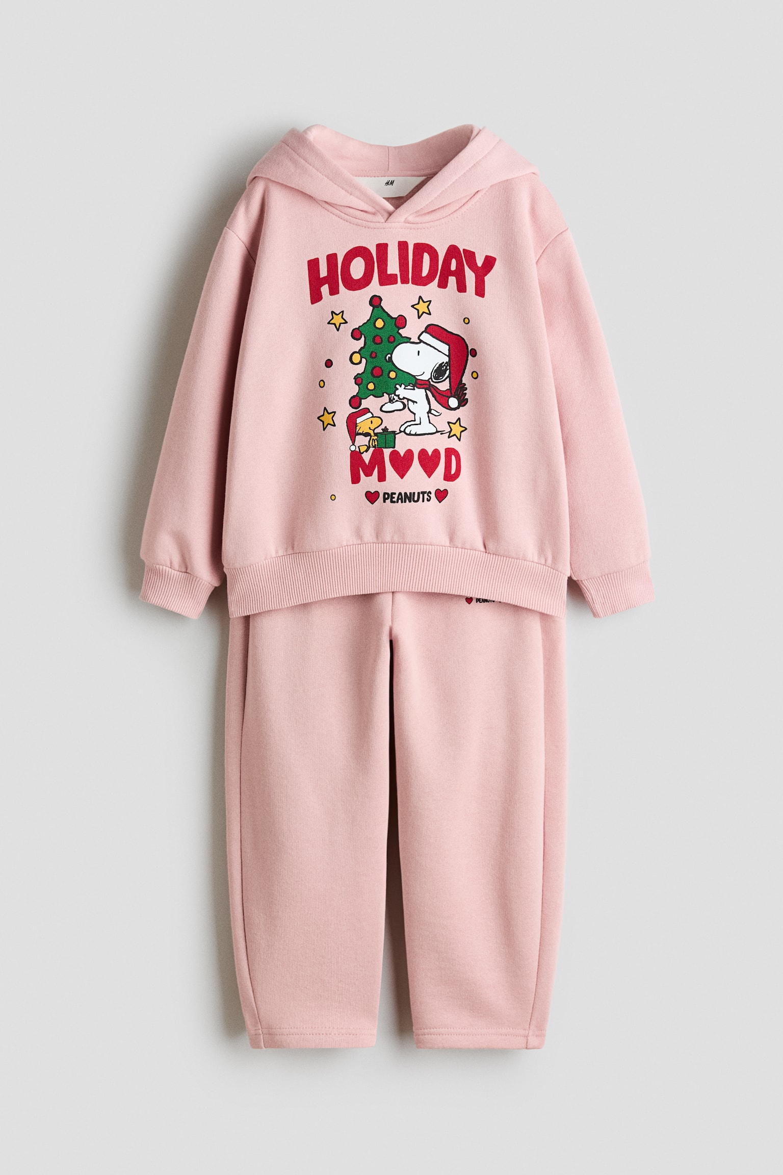 Ein hellrosa Fleece-Set ist von vorne zu sehen und besteht aus einem Hoodie mit langen Ãrmeln und einer dazu passenden Trainingshose. Auf dem Hoodie ist ein weihnachtliches Motiv mit Snoopy, Woodstock, einem Weihnachtsbaum und dem Schriftzug âHOLIDAY MOODâ auf der Brust zu sehen. Die passende Trainingshose zeigt einen elastischen Bund und leicht zulaufende Beine.