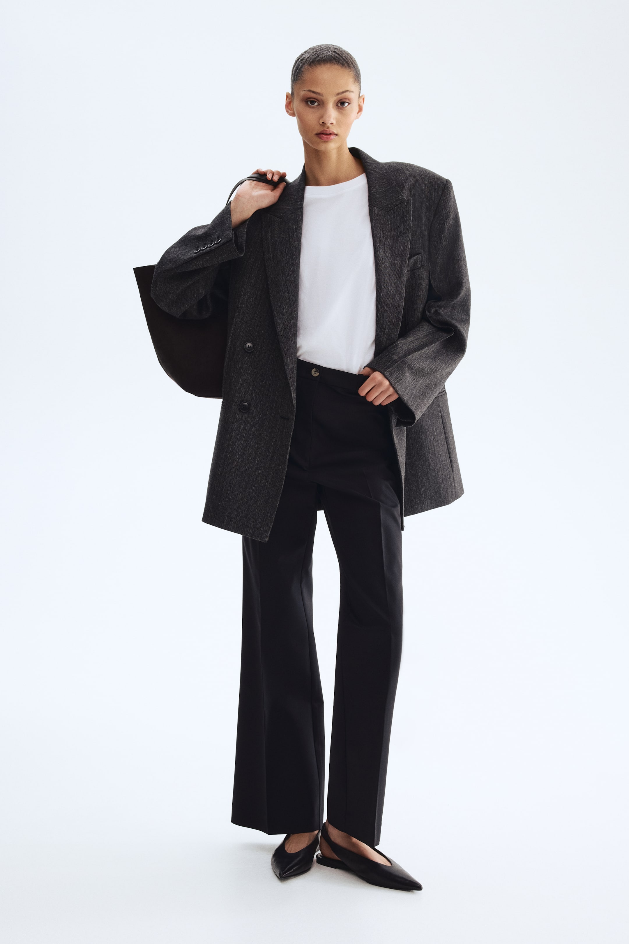 Ingrandisci l'immagine: Un blazer doppiopetto a righe sottili color grigio antracite viene indossato sopra una t-shirt girocollo bianca e pantaloni neri a gamba larga. Il blazer presenta ampi revers a lancia, tasche a pattina e tre bottoni su ciascun polsino, mentre delle ballerine slingback nere a punta completano l'insieme.