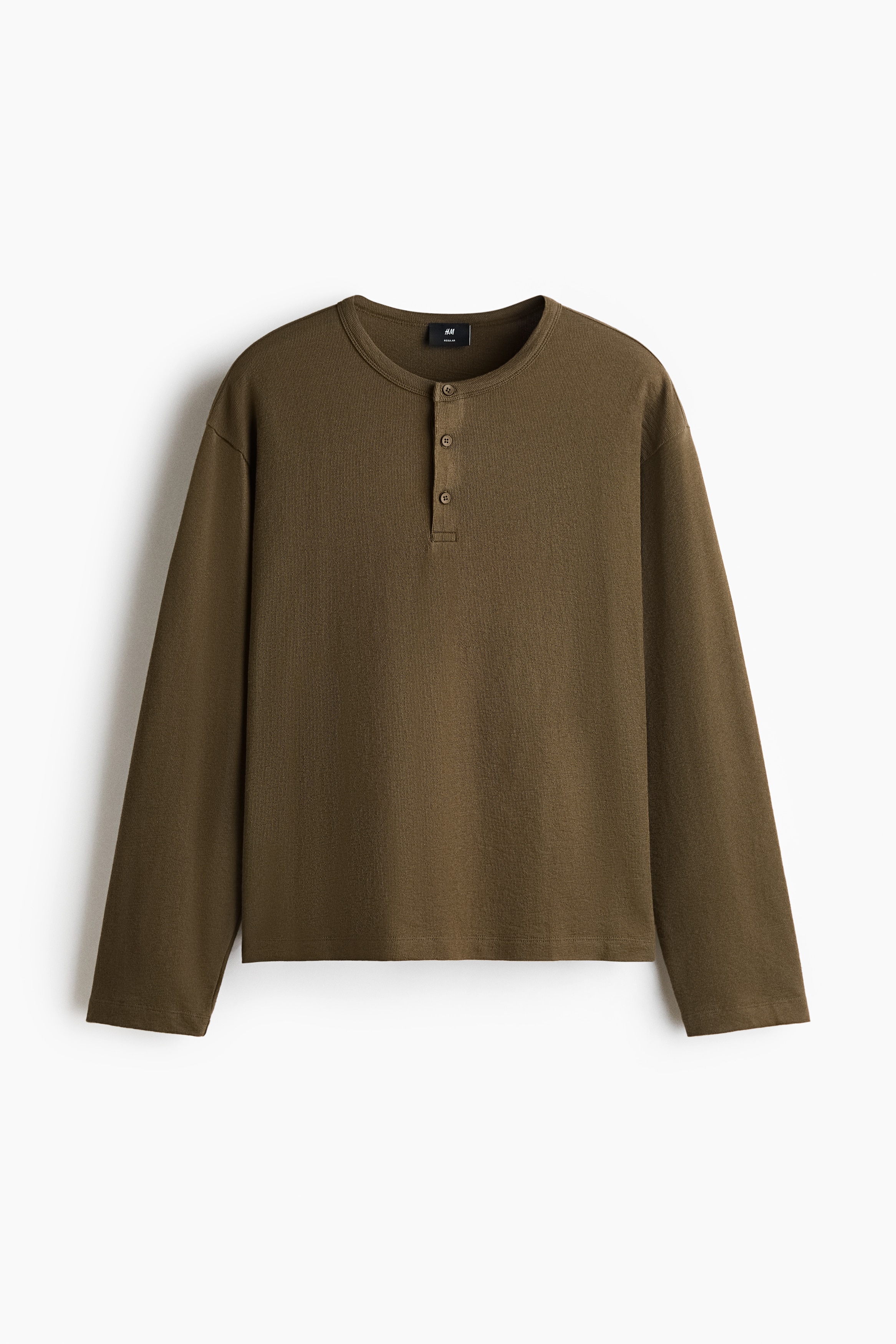 The model is wearing a long-sleeve henley shirt in olive green, featuring textured knit fabric and a partially unbuttoned collar. The relaxed-fit shirt is styled slightly tucked into black trousers with a black belt.