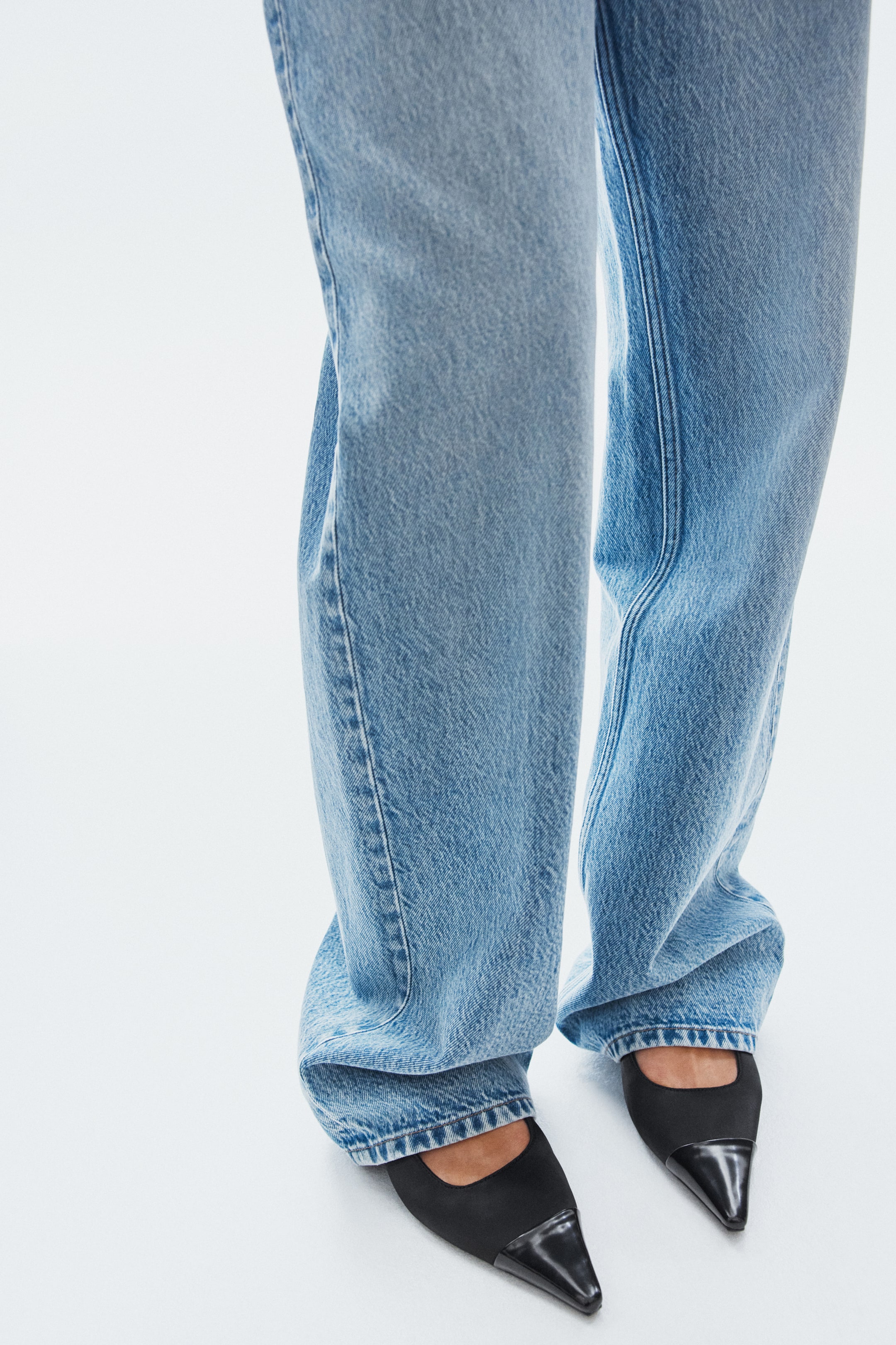 Nagyobb kép megtekintése: A modellen fekete szatén slingback cipők láthatók hegyes orral és fényes, bevont orr-résszel. Világoskék, bő szárú farmernadrágok omlanak a bokákra, és a cipők tetején nyugszanak.