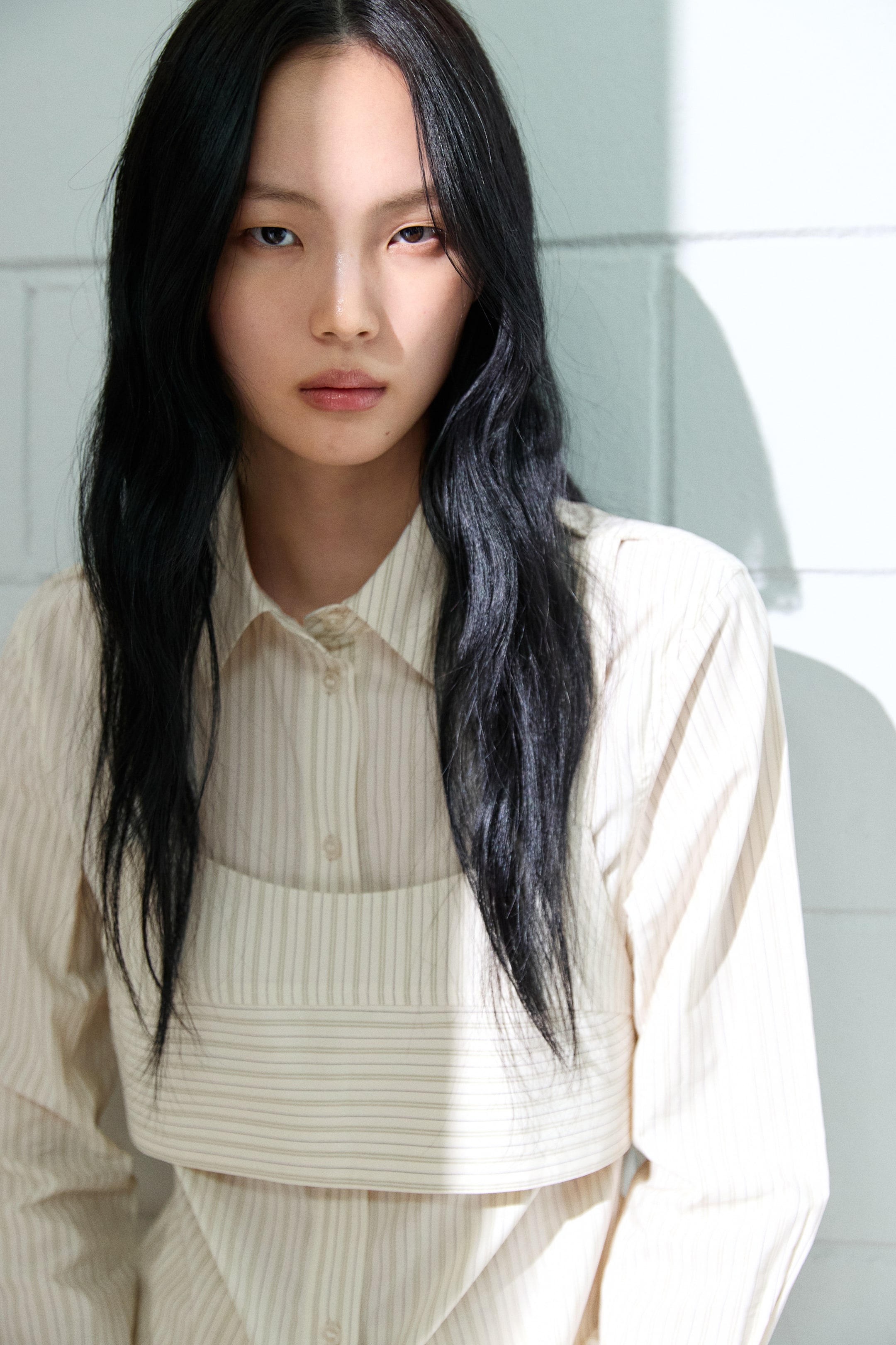 拡大画像を表示: 薄いベージュの襟付きシャツが紹介されています。細い縦縞とボタン留めの前立てが特徴です。シャツの胸部と上腹部には、構造的な横縞のバンドが重ねられ、独特なデザインを生み出しています。