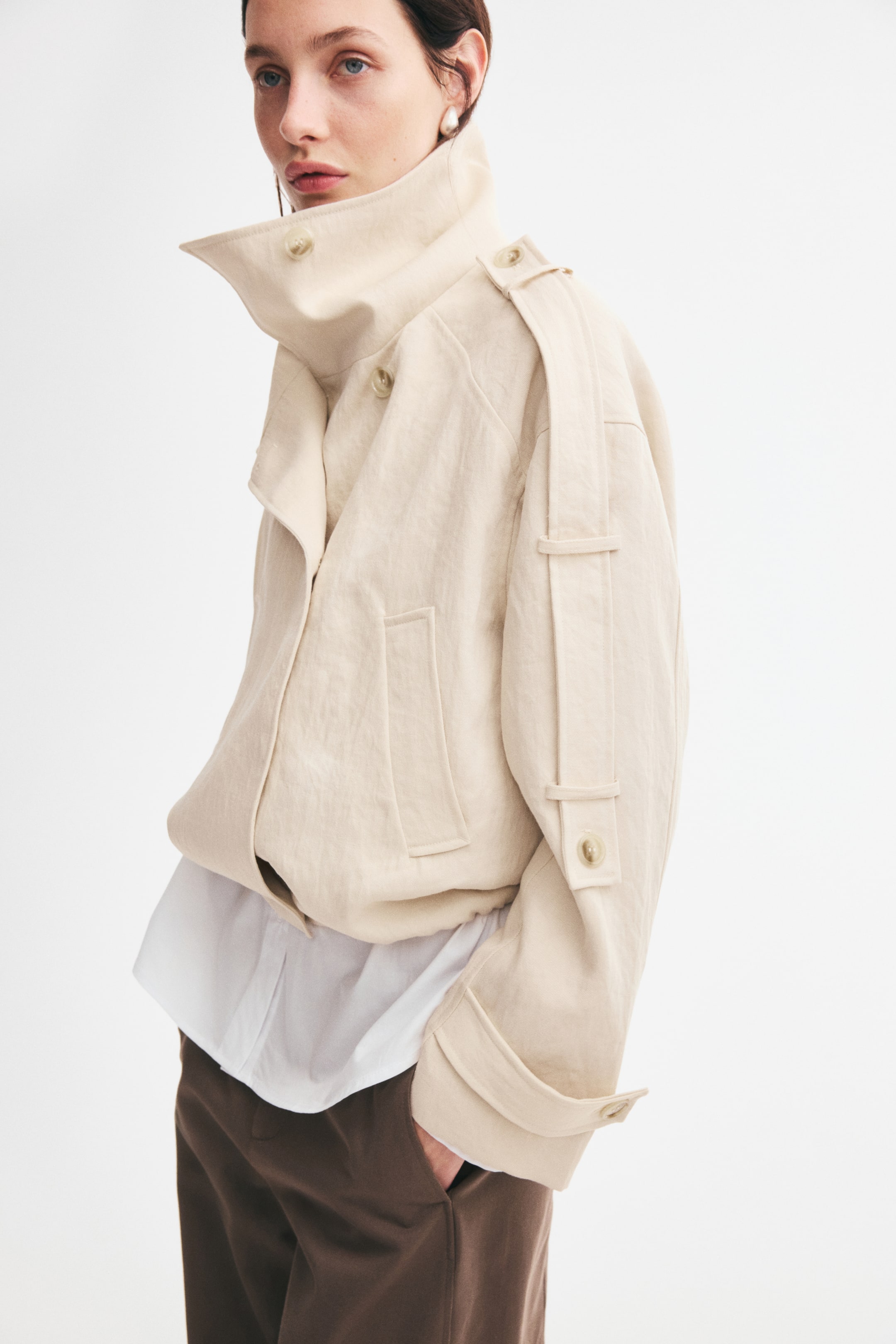 View larger image: The model is wearing a short, oversized light beige trench jacket with a high, structured funnel collar and decorative buttoned tabs along the shoulder and sleeve. Layered over a white shirt and brown trousers, it features a slightly gathered bubble hem and a welt pocket.
