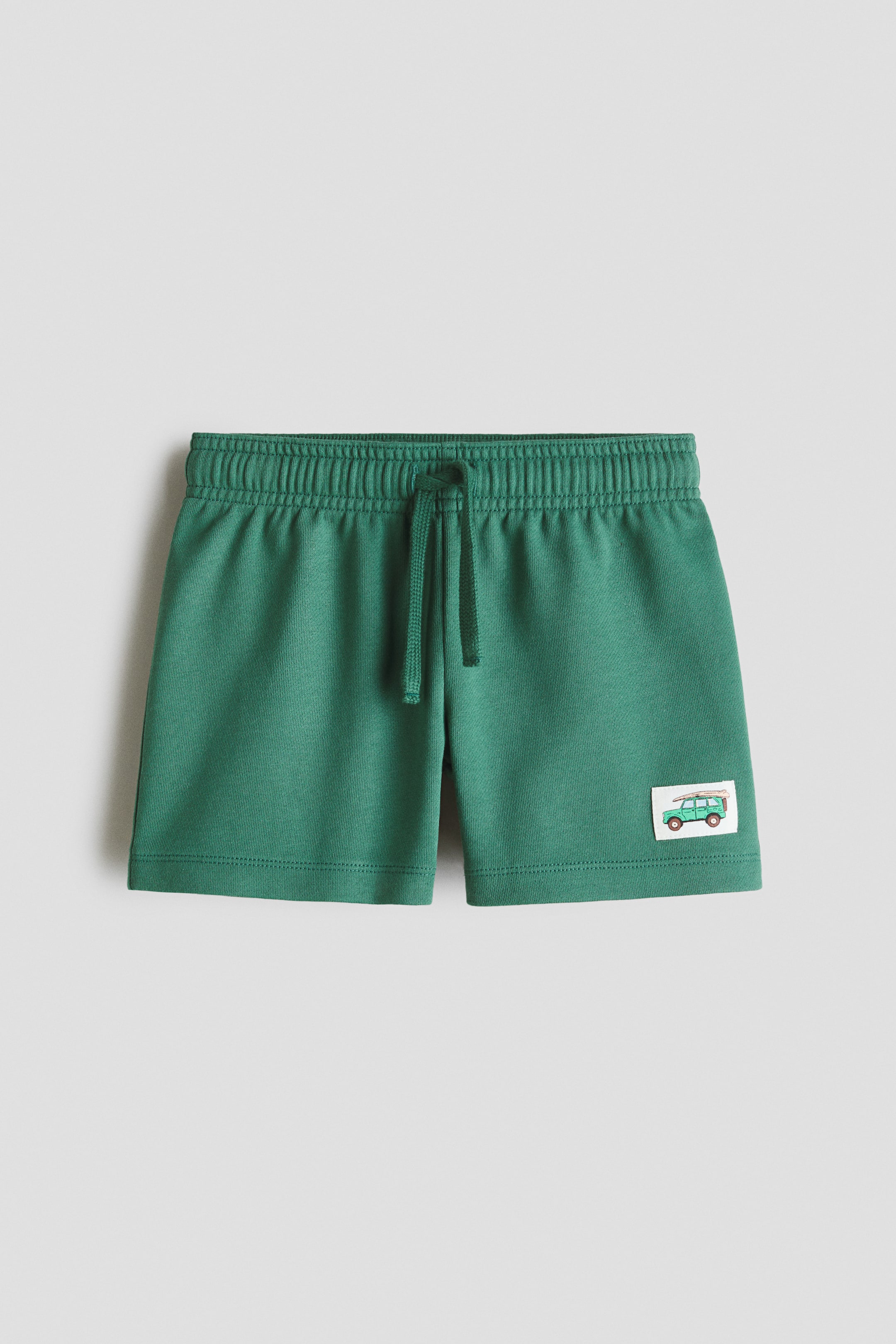 View larger image: A pair of green shorts is displayed from the front, showcasing an elasticated waistband with a green drawstring tie. A small rectangular patch is visible on the lower right leg, illustrating a green off-road vehicle carrying a surfboard.