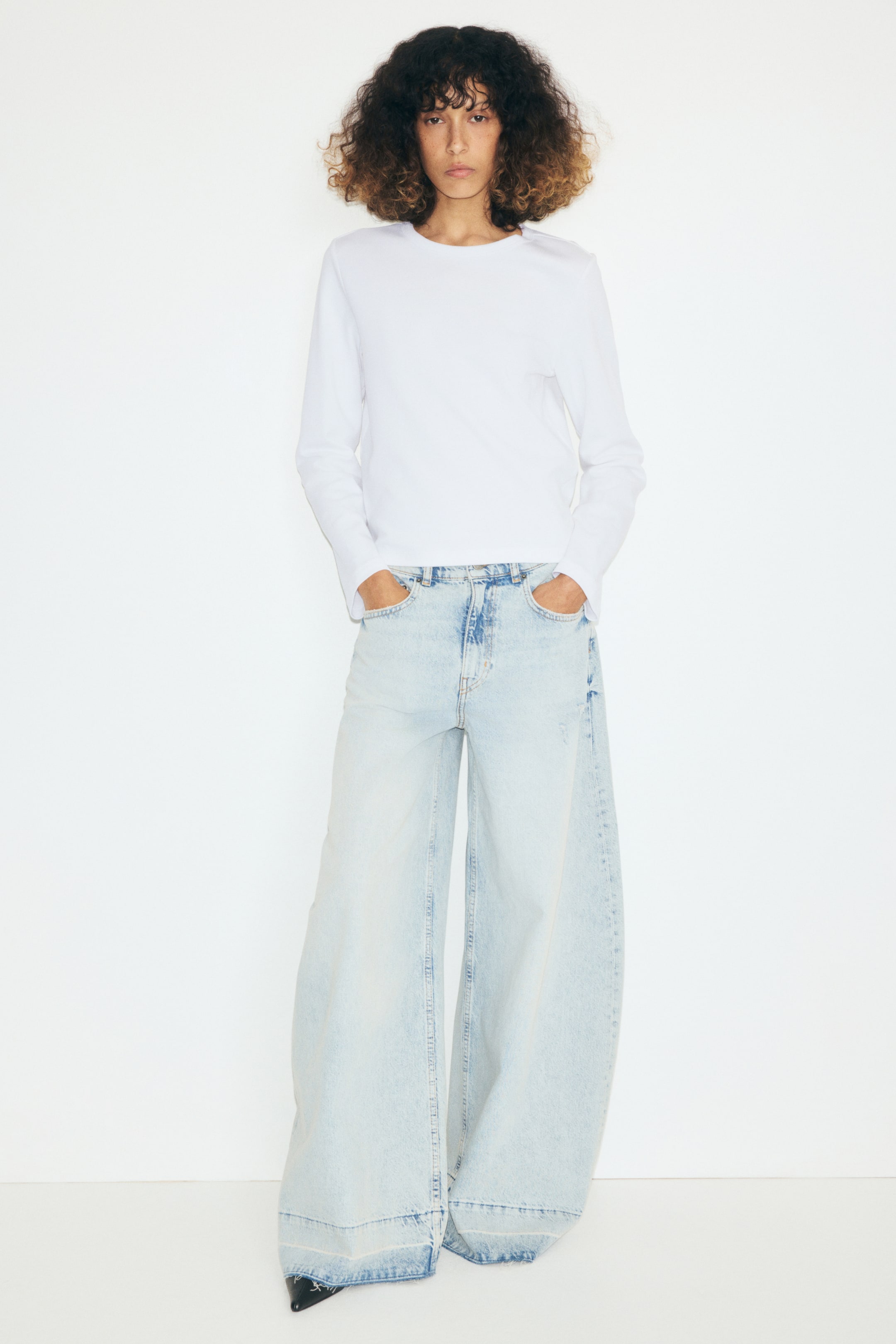 View larger image: The model is wearing a white top made from heavy jersey, featuring a round neckline with ribbed trim and long sleeves. The top is paired with light-wash wide-leg jeans, with the model’s hands resting in the front pockets.