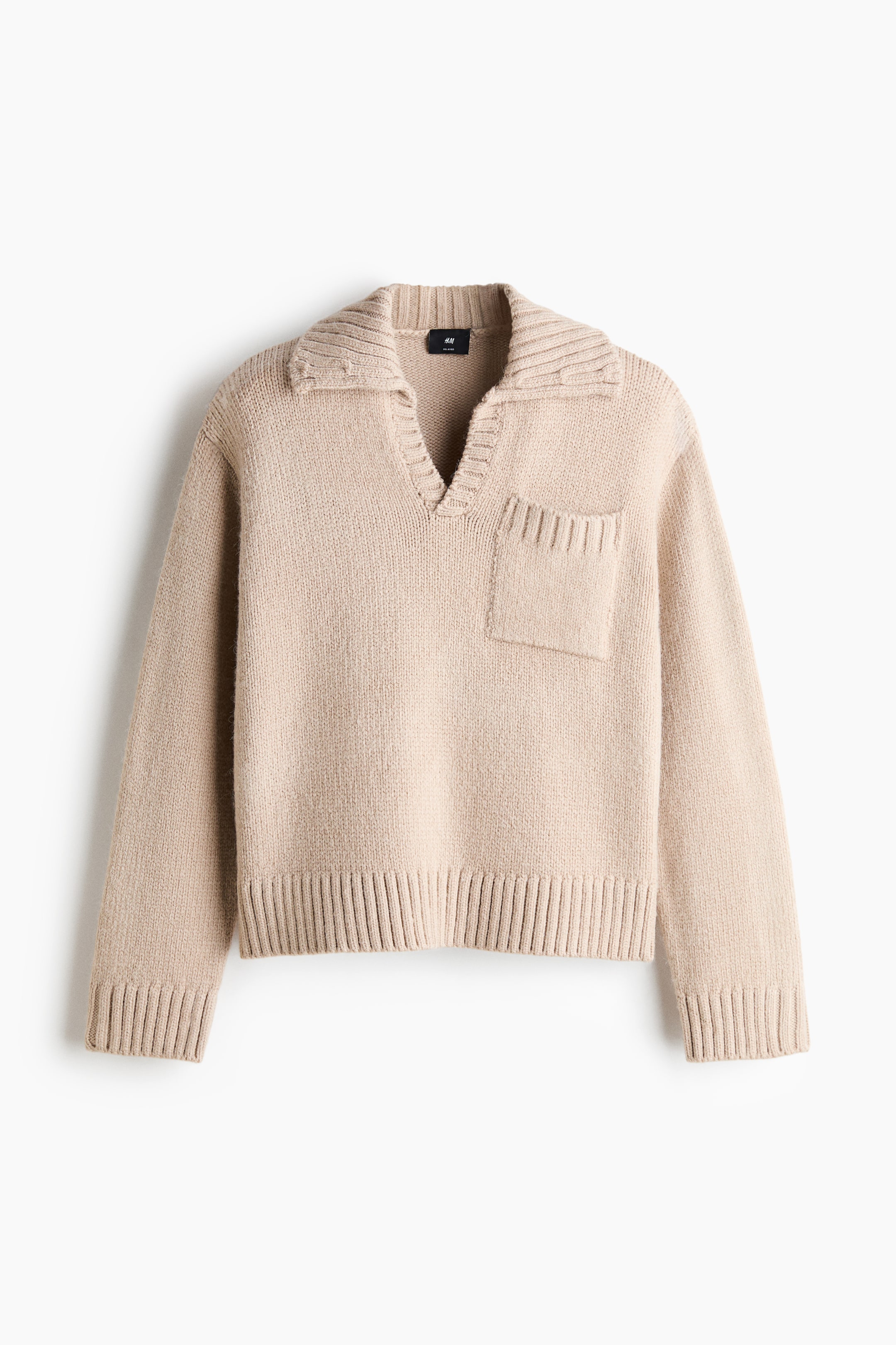 Visa större bild: En ljusbeige stickad tröja är vänd framåt och visar upp en ribbad krage med en öppen V-ringning. Den har en enkel påsydd ficka med ribbad kant på vänster bröst, långa ärmar med breda ribbade muddar och en matchande ribbad nederkant.