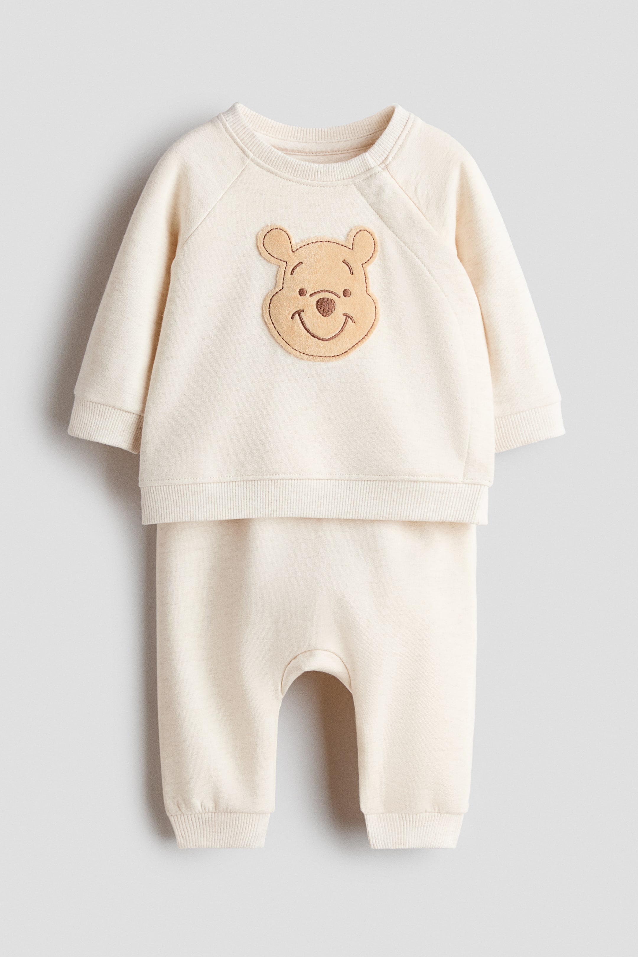 View larger image: A cream two-piece baby outfit is laid flat, consisting of a long-sleeved sweatshirt with a crew neck, raglan sleeves, ribbed cuffs, and hem, and a Winnie the Pooh face appliqué on the chest. It is paired with matching long pants, featuring a dropped crotch and ribbed ankle cuffs.