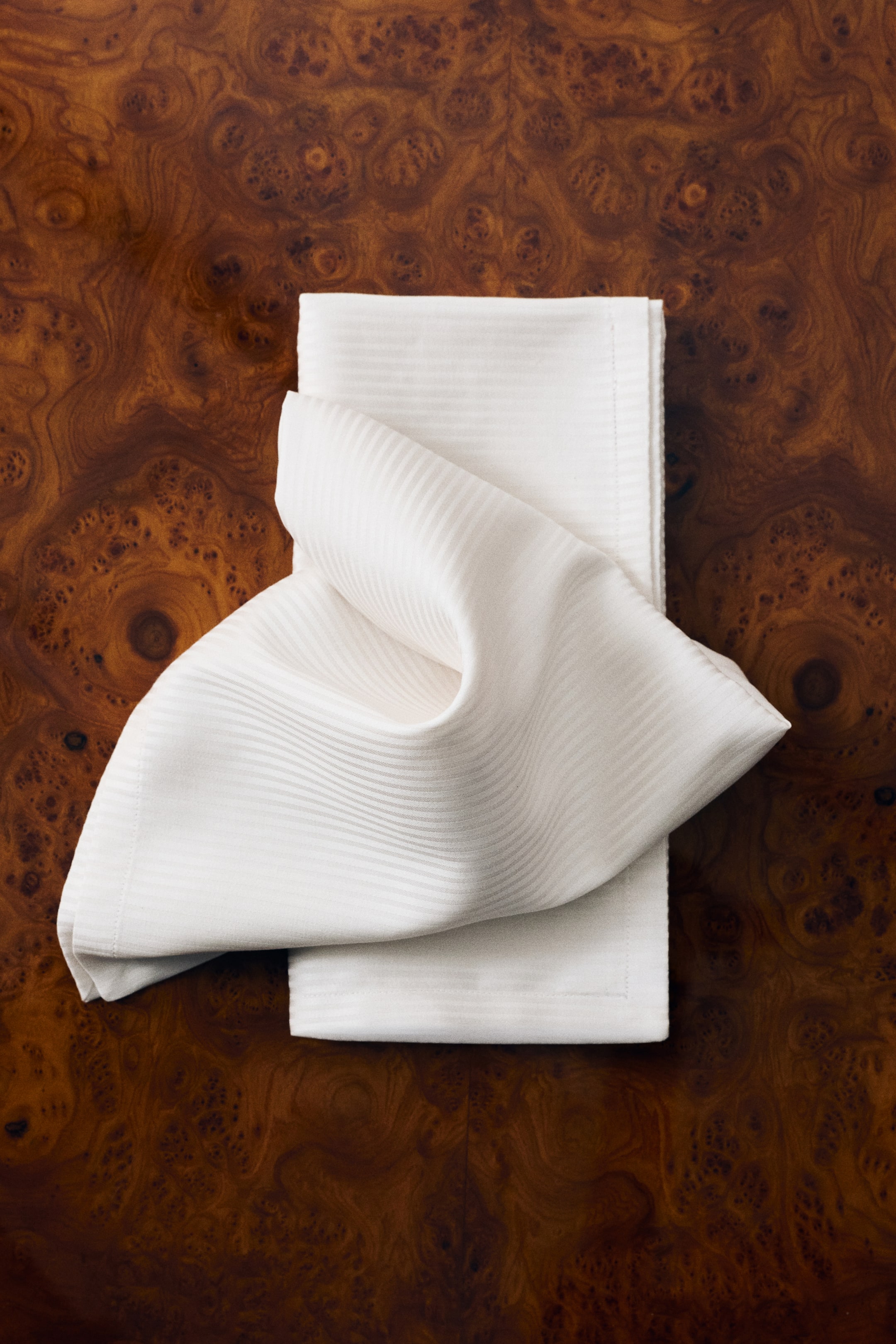 View larger image: Two off-white cotton-blend napkins are shown, with one folded flat in a rectangular shape and the other draped loosely on top in a soft, wavy fold. The napkins display a subtle, tone-on-tone striped woven pattern and a folded hem along the edges.
