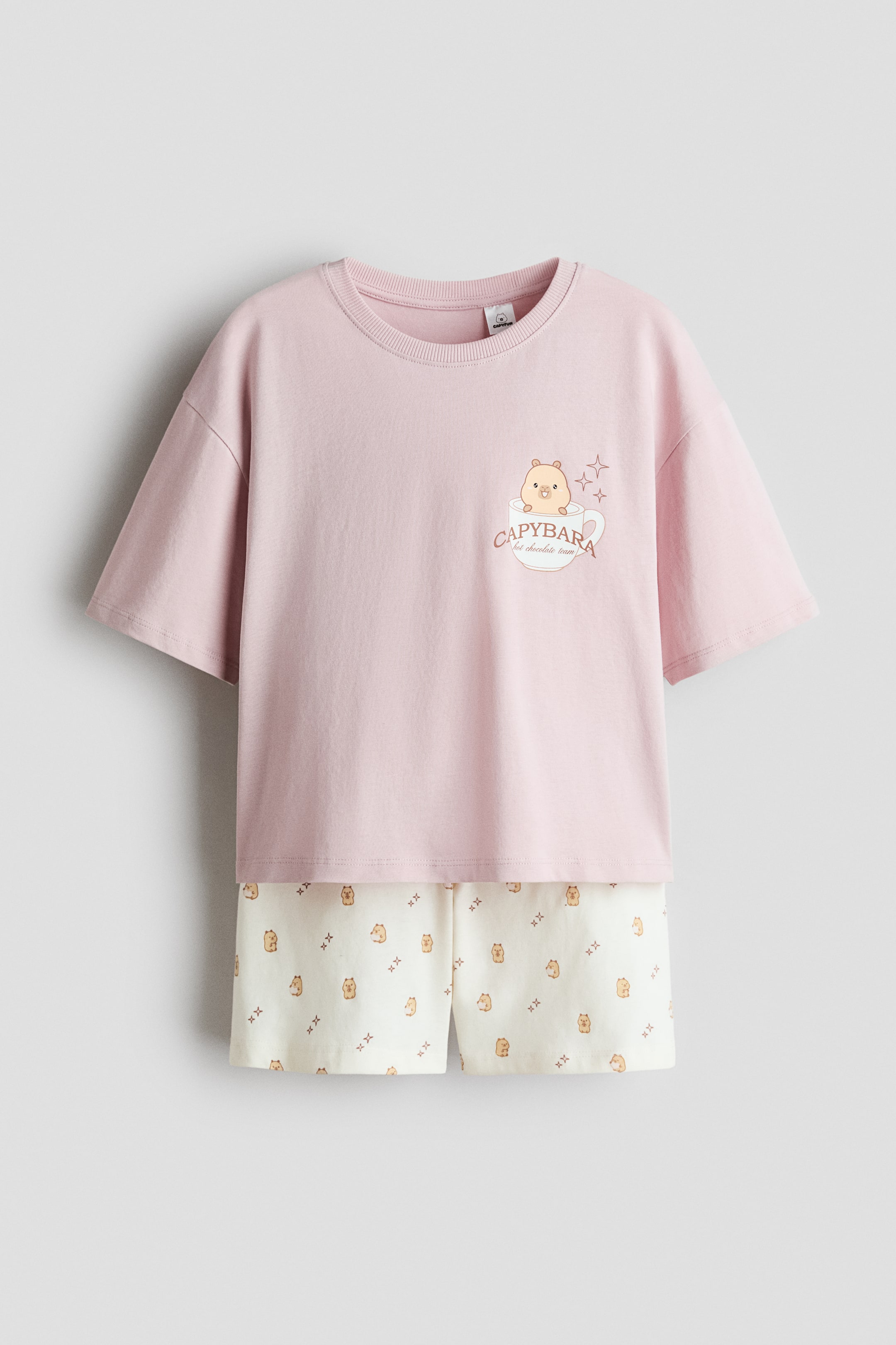 Agrandir l'image: Un t-shirt rose à manches courtes avec un col rond côtelé arbore un graphique d'un capybara dans une tasse blanche, accompagné du texte "équipe chocolat chaud capybara" et d'étoiles. Un short crème assorti présente un motif répétitif de petites figures de capybara et de formes croisées.