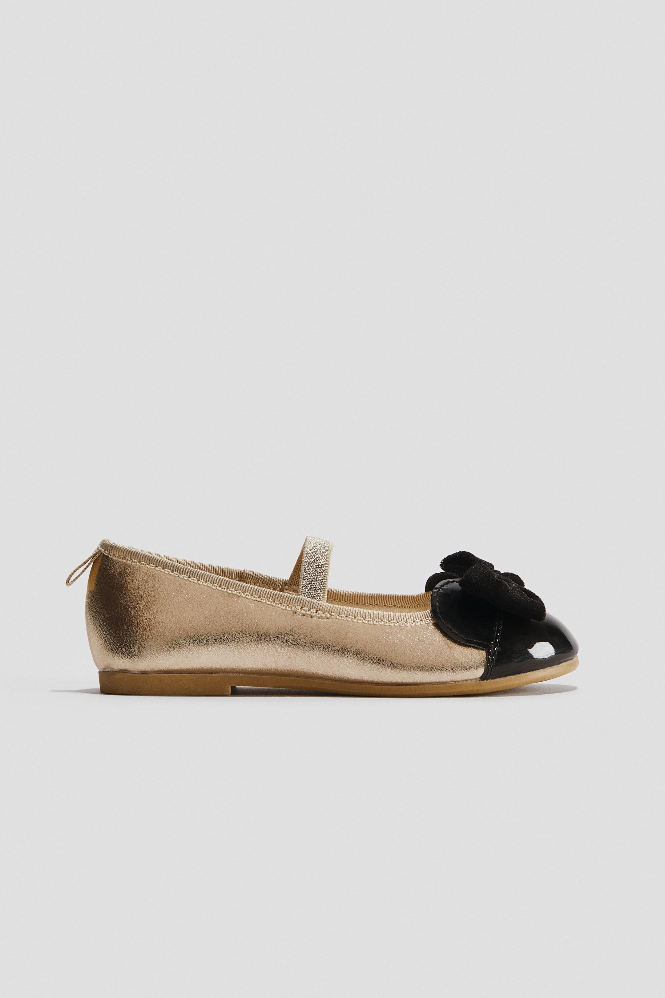 View larger image: A metallic gold Mary Jane flat showcases a black patent toe cap adorned with a black velvet bow. A glittering gold strap extends across the instep.
