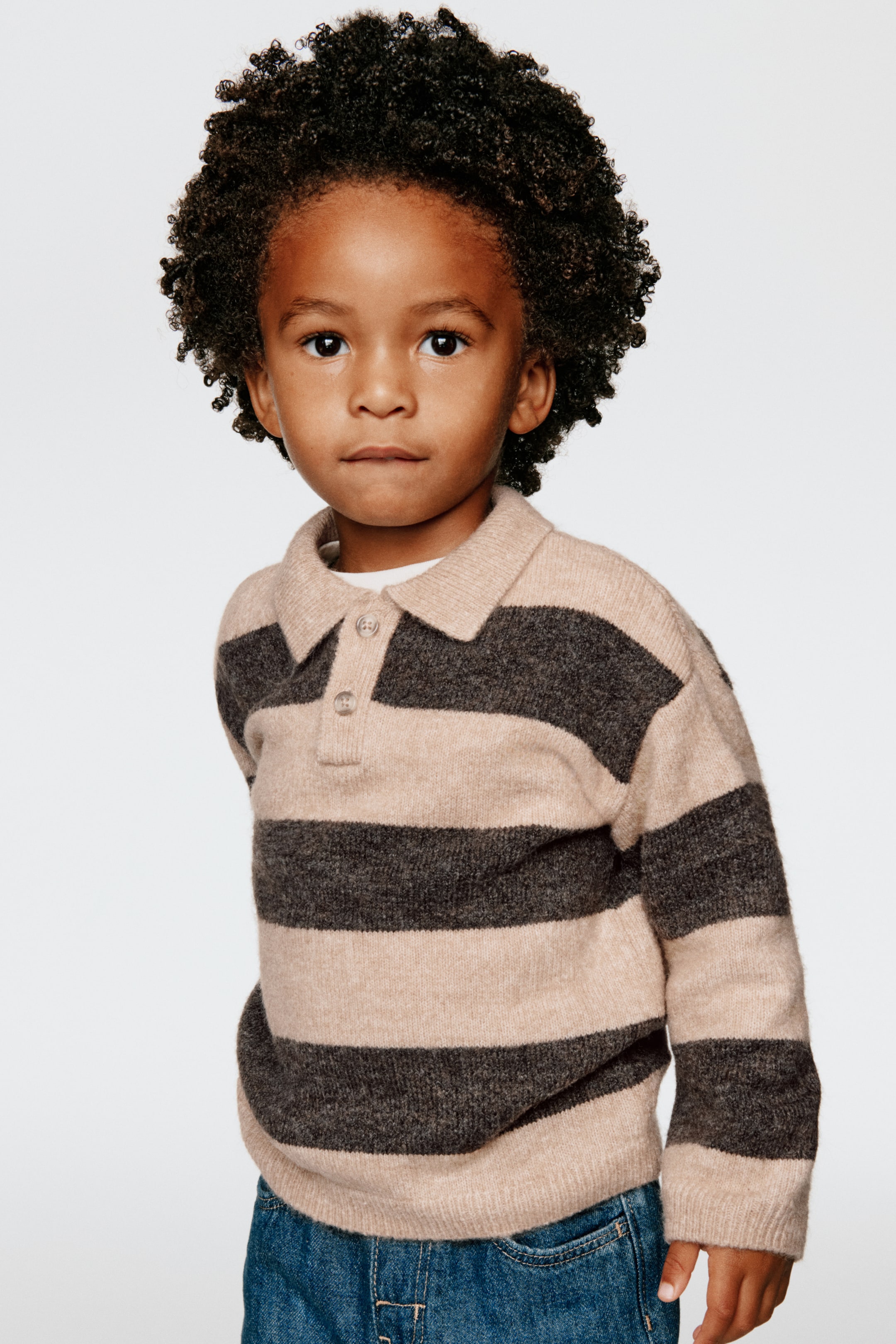 Ver imagen más grande: Se muestra por delante un jersey de punto tipo polo con rayas horizontales beige y gris oscuro, con cuello vuelto, tapeta de dos botones y hombros caídos. El jersey, superpuesto sobre una camiseta blanca, cuenta con puños acanalados y bajo acanalado.