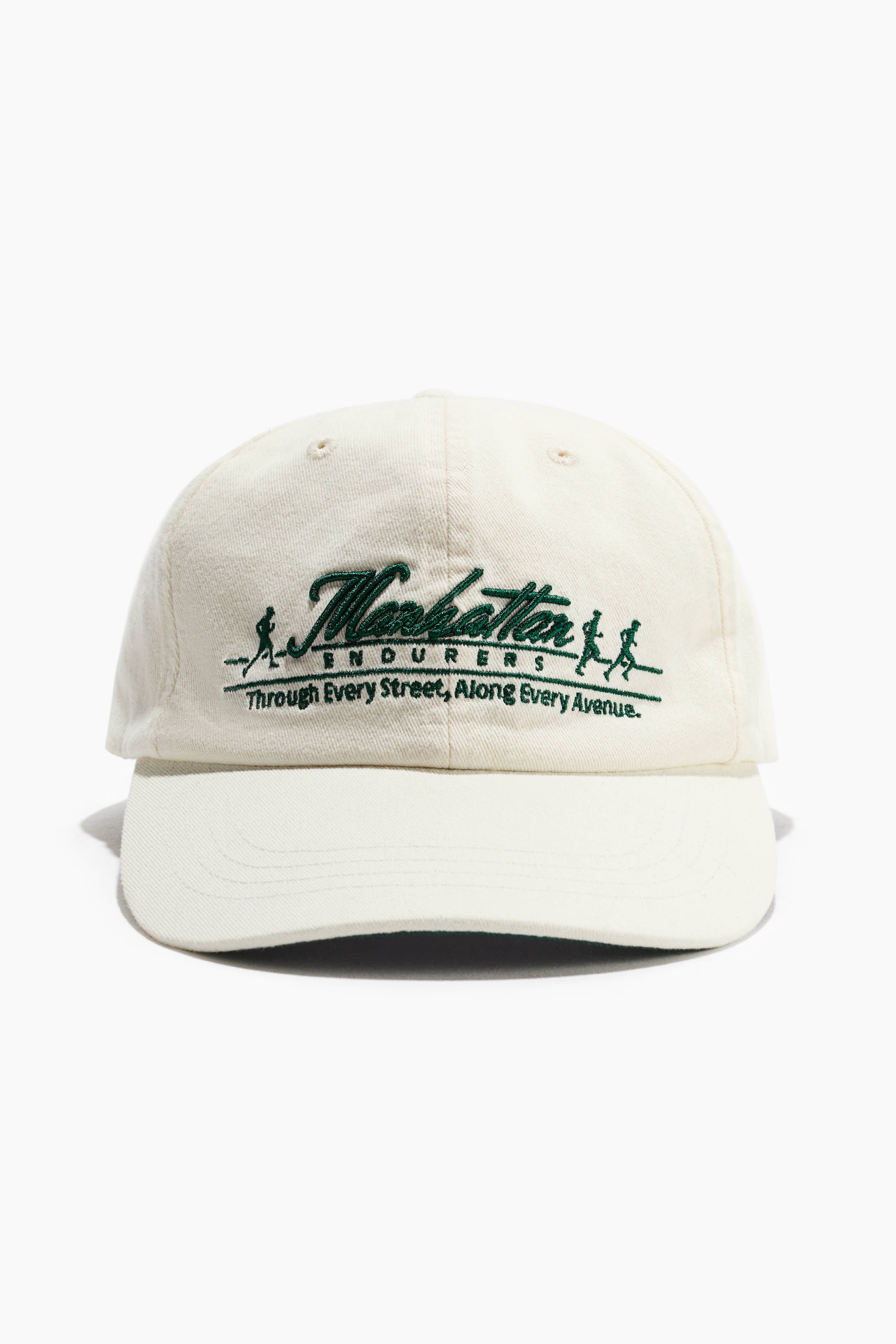 Agrandir l'image: Une casquette en sergé de coton de couleur crème est présentée de face, mettant en valeur une visière incurvée et des œillets brodés sur la calotte. L’avant affiche un motif brodé vert avec les mots « Manhattan ENDURERS », accompagnés de silhouettes de coureurs et de texte.
