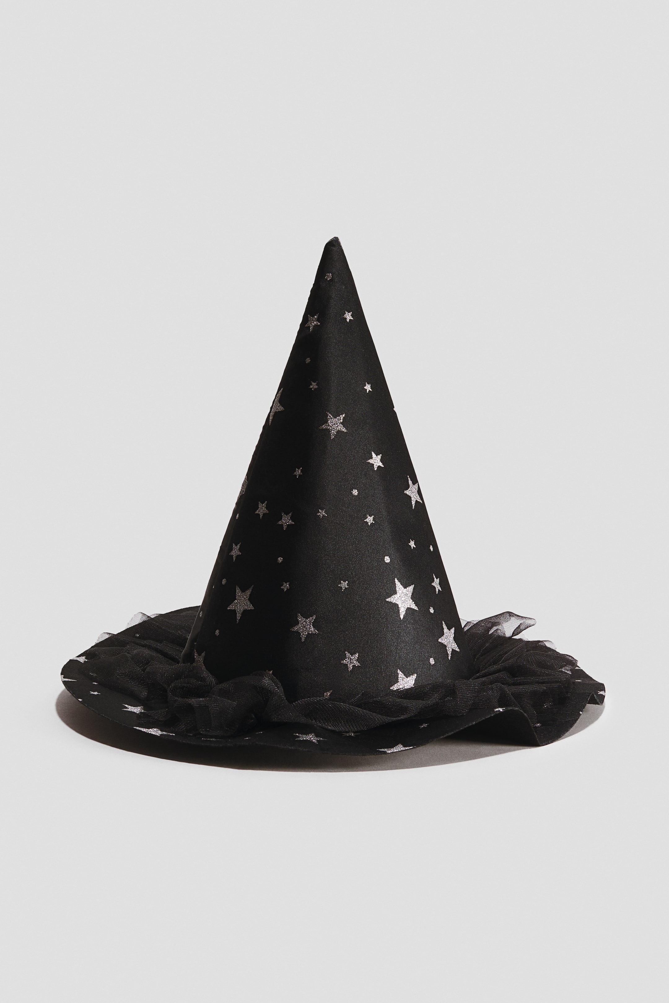 View larger image: A black conical hat features silver glitter stars in various sizes. A ruffled black tulle trim decorates the base of the cone, resting on the wide brim.