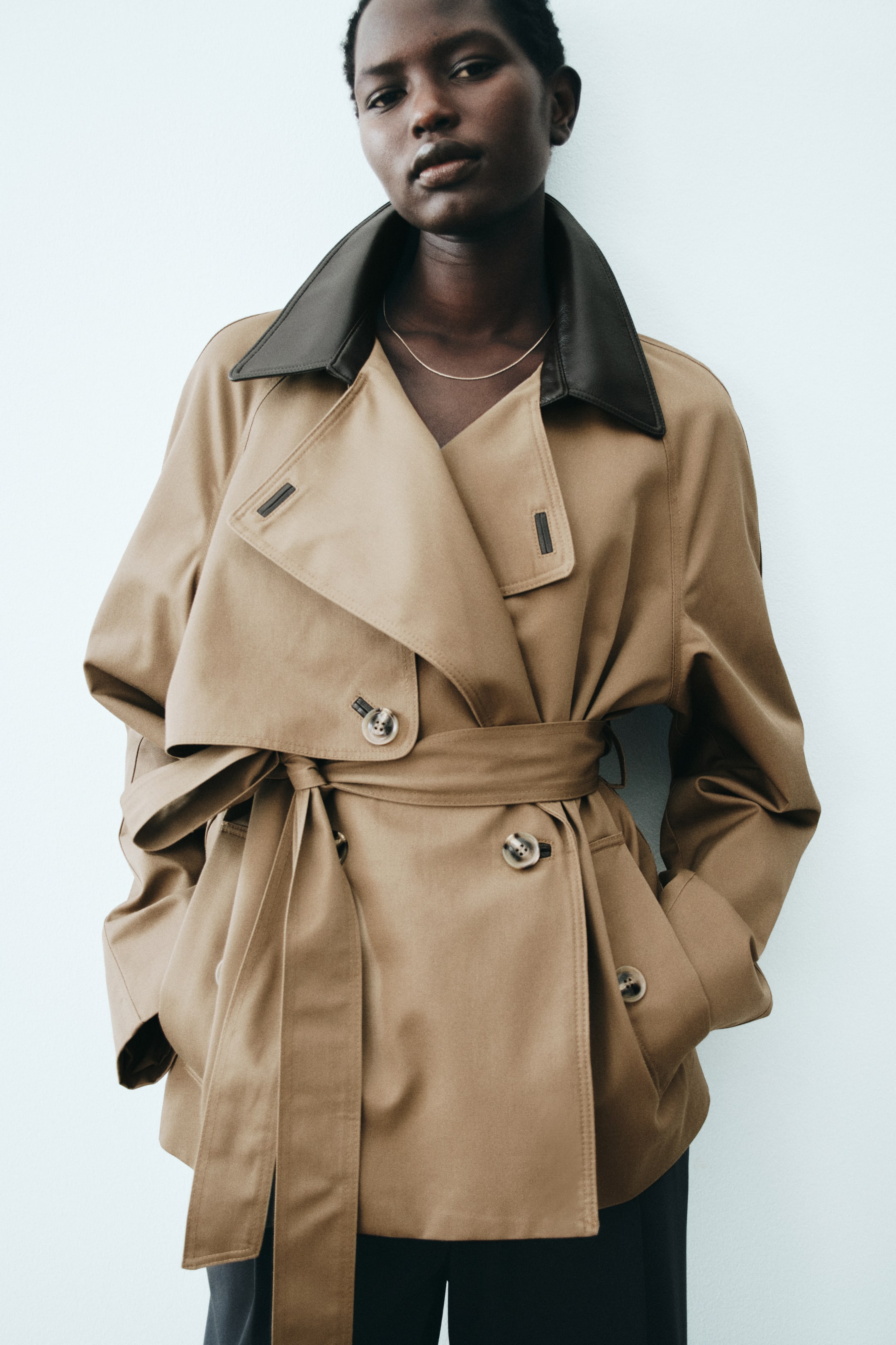 Ingrandisci l'immagine: Un trench coat color cammello è indossato, caratterizzato da un colletto a contrasto marrone scuro, un davanti doppiopetto con bottoni marroni e una pattina antipioggia sul petto destro. Una cintura in tessuto con nodo automatico stringe la vita, mentre le mani riposano nelle sue tasche a fessura.