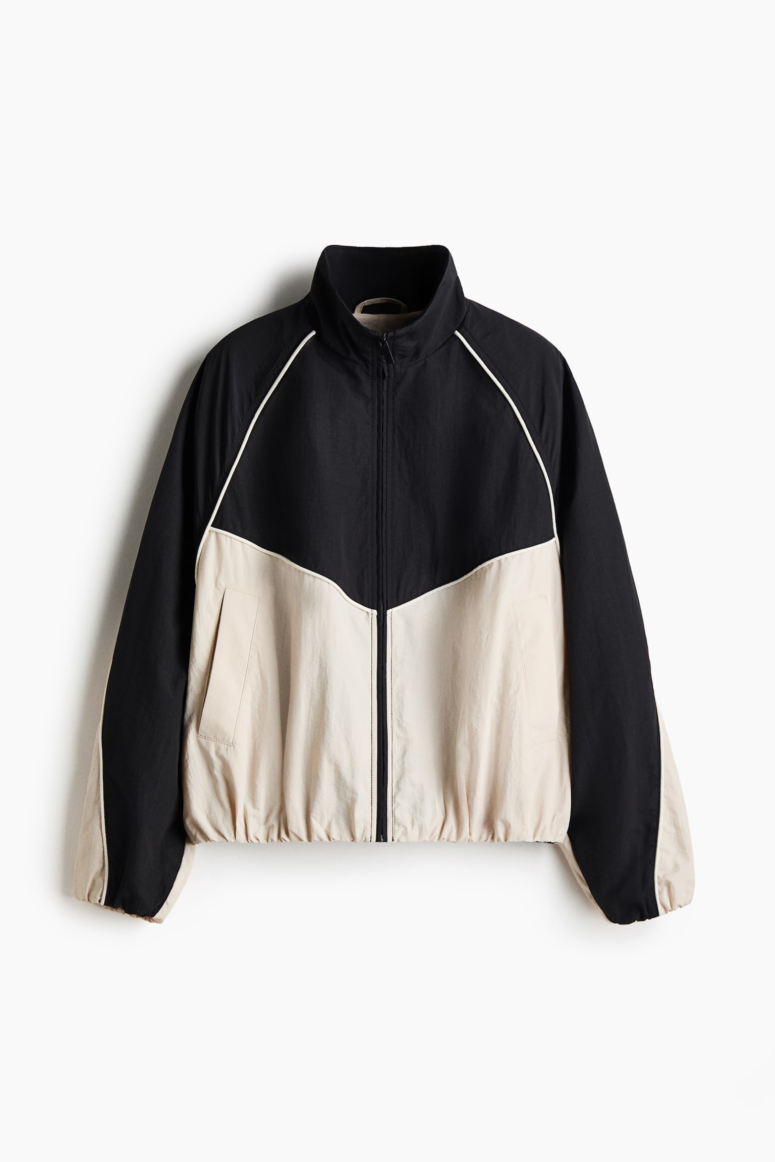 This front-facing colour-blocked jacket features a high stand-up collar, a central zip fastening, and an elasticated hem and cuffs. The design showcases a black upper section and outer sleeves contrasted with a cream-coloured lower body and inner sleeves, with thin white piping highlighting the chevron chest seam and raglan shoulders.