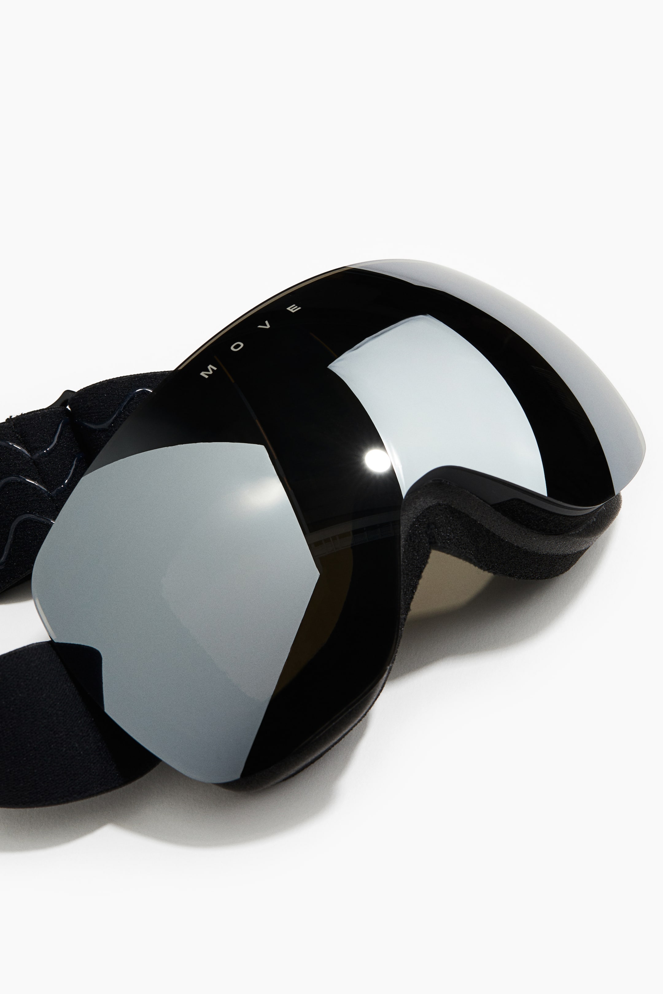 Visa större bild: Svarta skidglasögon har en stor, böjd silverfärgad spegellins och en svart ram med "MOVE"-märkning synlig på toppen. Svart skumvaddering täcker insidan, och ett svart elastiskt band sträcker sig från vänster sida.