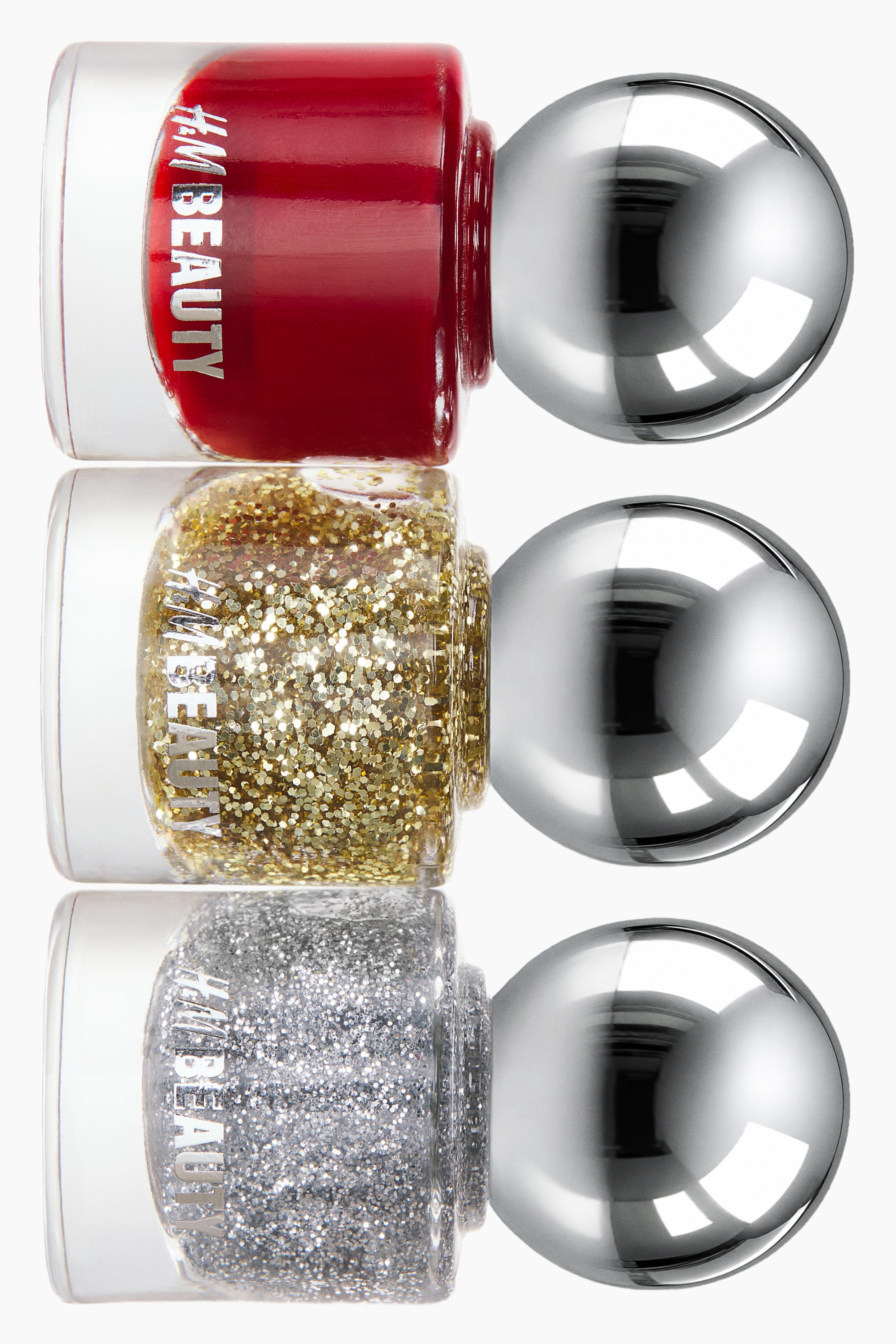 View larger image: Three nail polish bottles are positioned horizontally in a vertical stack, showing cylindrical glass bodies and large, reflective, spherical silver caps. The bottles feature a solid red polish at the top, a gold glitter polish in the middle, and a silver glitter polish at the bottom, with each displaying the "H&M BEAUTY" logo printed on the side.