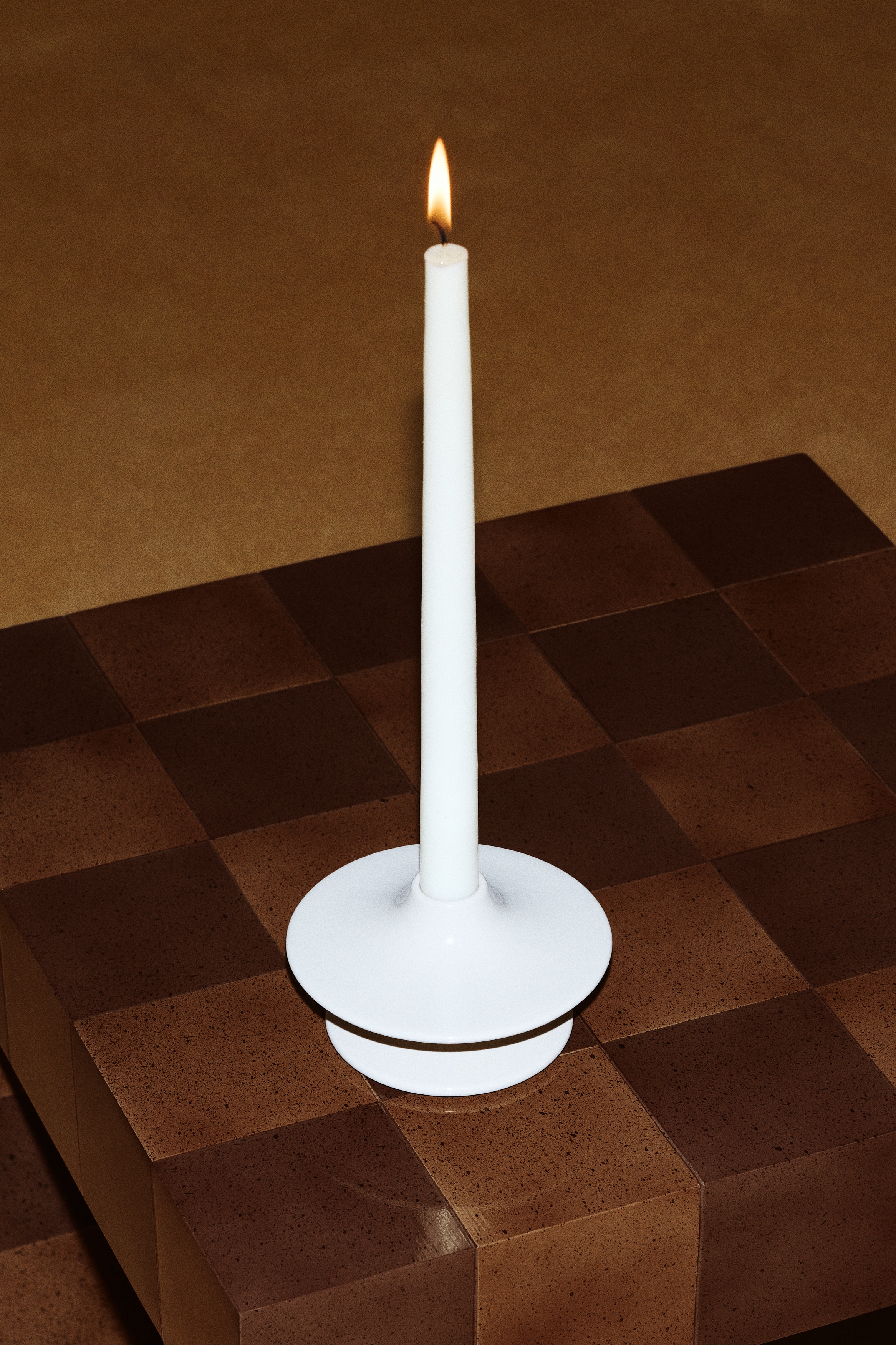 A light beige ceramic candlestick is holding an off-white tapered candle, featuring a circular base and a wide, flat top with a subtly curved, upward-sloping edge.