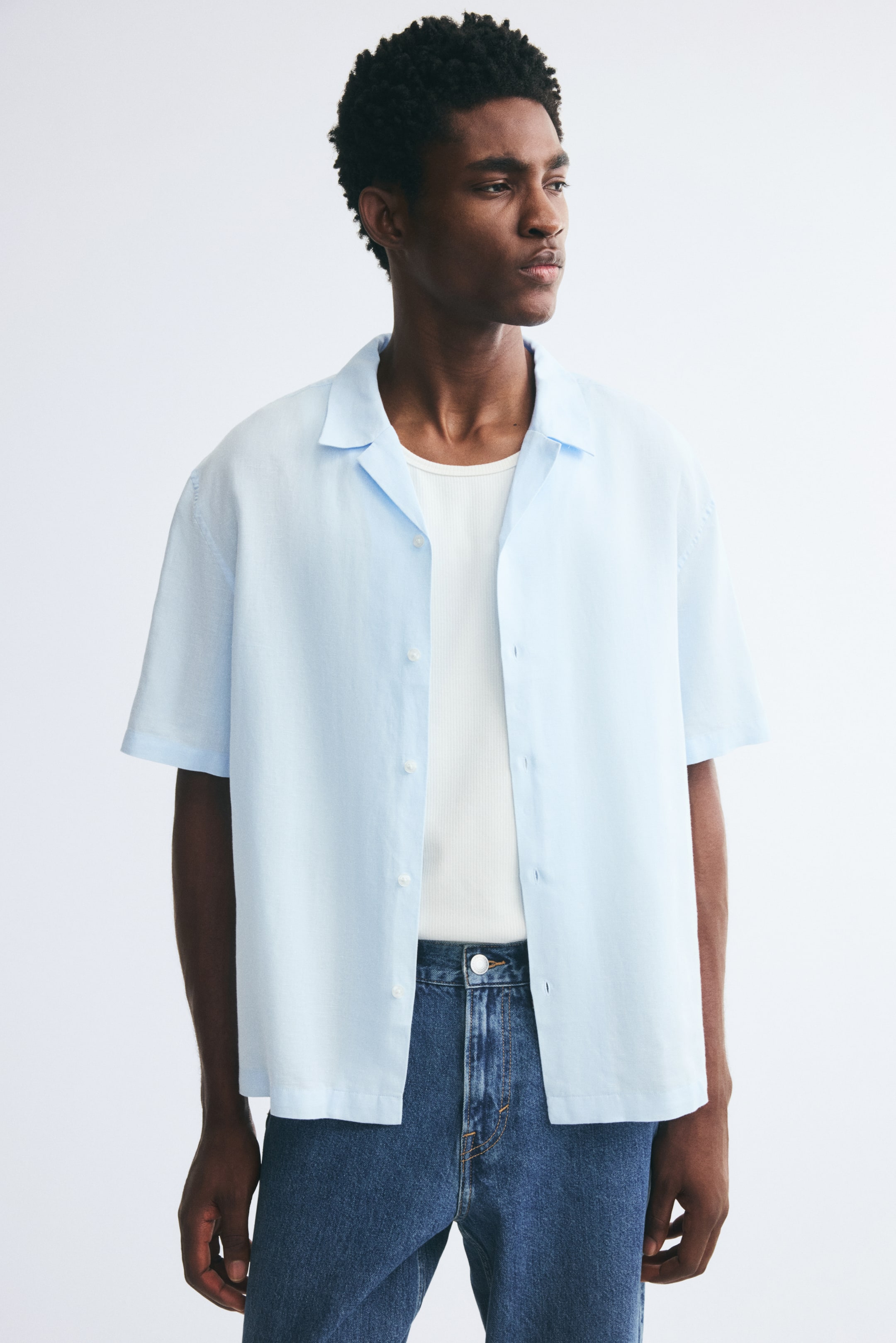 Xem ảnh lớn hơn: Một chiếc áo sơ mi tay ngắn màu xanh nhạt, làm từ vải dệt cotton và linen có bề mặt nổi, được người mẫu mặc mở cúc. Áo có phom dáng thoải mái, cổ resort và lai áo cắt thẳng, được phối bên ngoài áo hai dây trắng gân cùng quần jeans xanh.