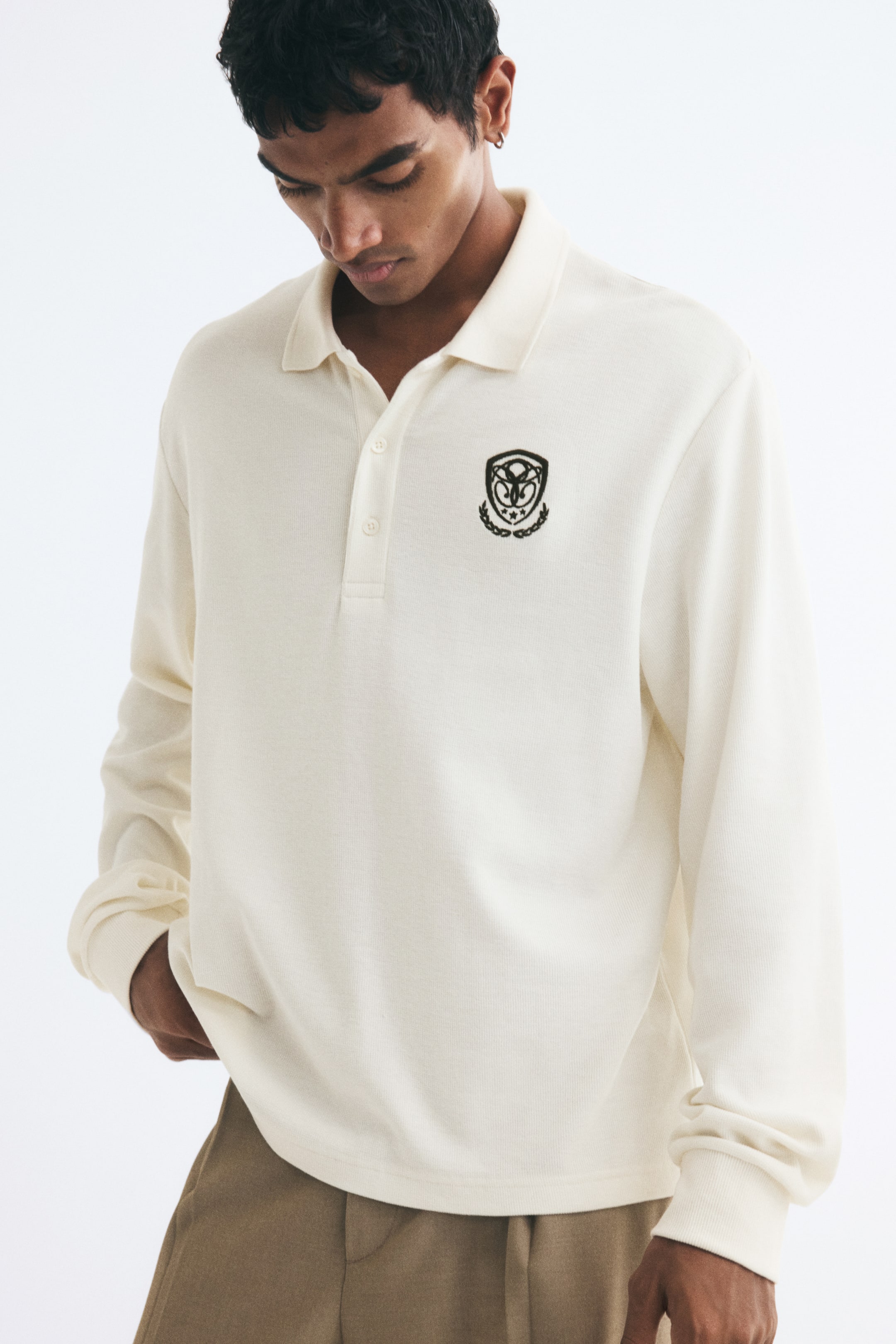 View larger image: A cream-coloured, long-sleeve polo shirt in a textured waffle knit is being worn by the model. The shirt features a ribbed collar, a button placket, ribbed cuffs, and a black embroidered crest on the chest. The model is pairing the shirt with beige trousers, placing one hand in a pocket.
