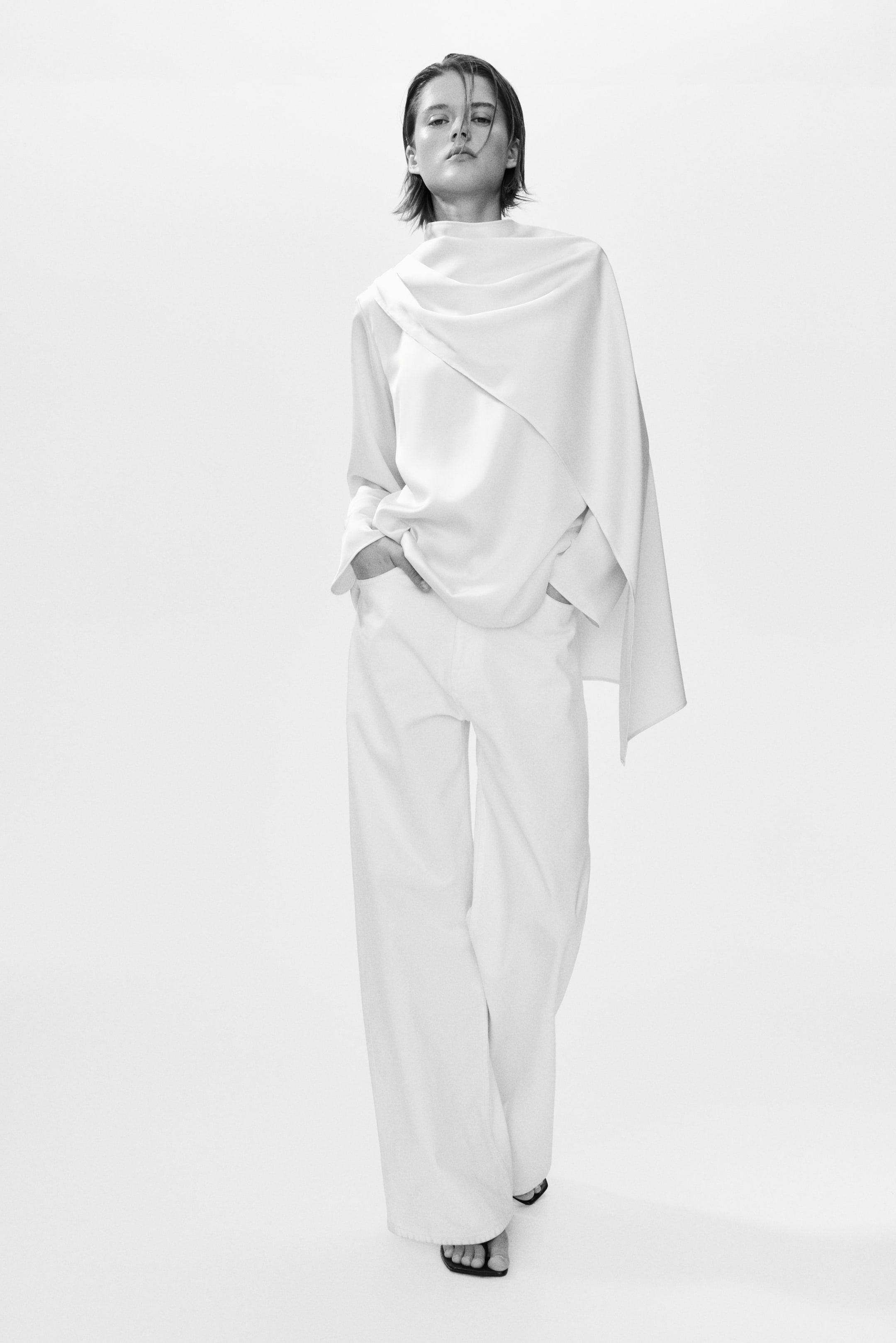 Agrandir l'image: Une blouse blanche présente un col montant et un drapé asymétrique s'étendant de l'épaule gauche, créant une silhouette fluide. Un pantalon blanc à jambes larges complète le look, mettant en valeur une coupe décontractée.