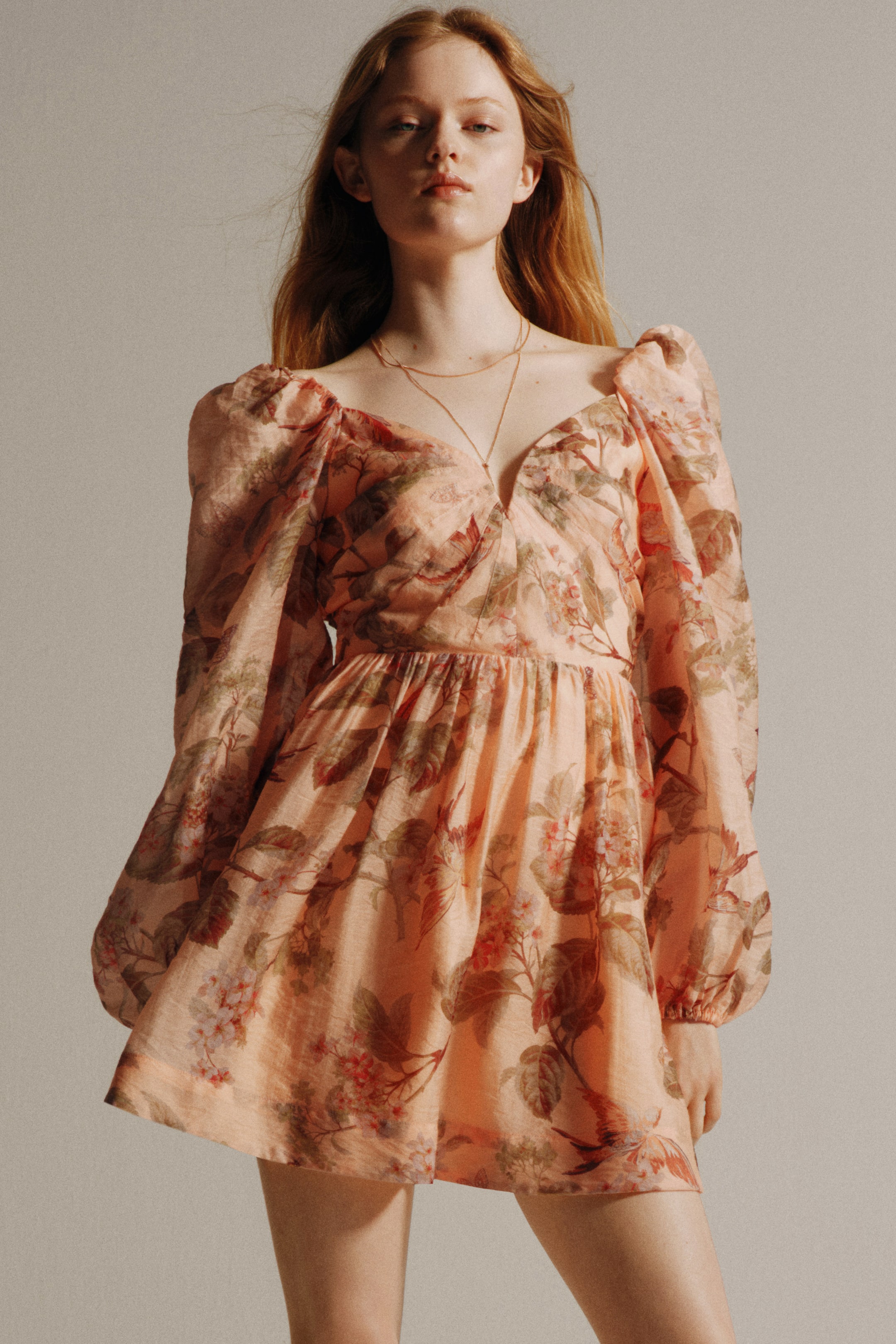 이미지 확대 보기: 이 드레스는 복숭아 색상의 원단에 꽃무늬가 돋보이며, 스위트하트 네크라인과 긴 퍼프 소매가 특징입니다. 허리 부분에서 주름이 잡혀 짧고 플레어 스커트로 이어집니다.