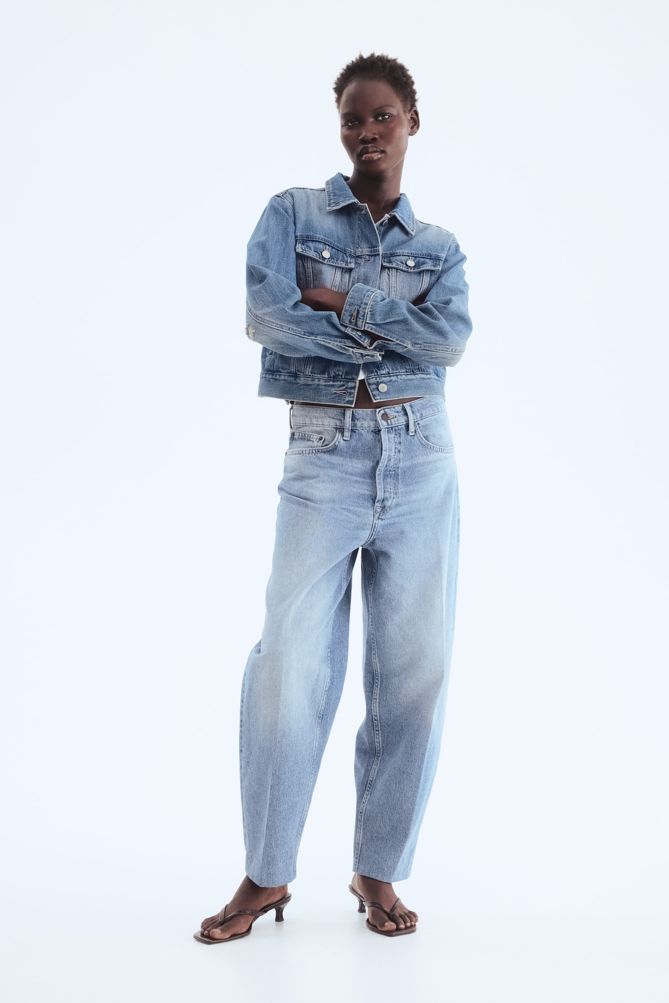 Größeres Bild anzeigen: Eine hellblaue Jeansjacke wird von vorne gezeigt, mit klassischem Kragen, Knopfverschluss vorne und Brusttaschen mit Klappe, kombiniert mit hoch taillierten, weit geschnittenen Jeanshosen und braunen Zehenstegsandaletten mit Kitten-Heel.