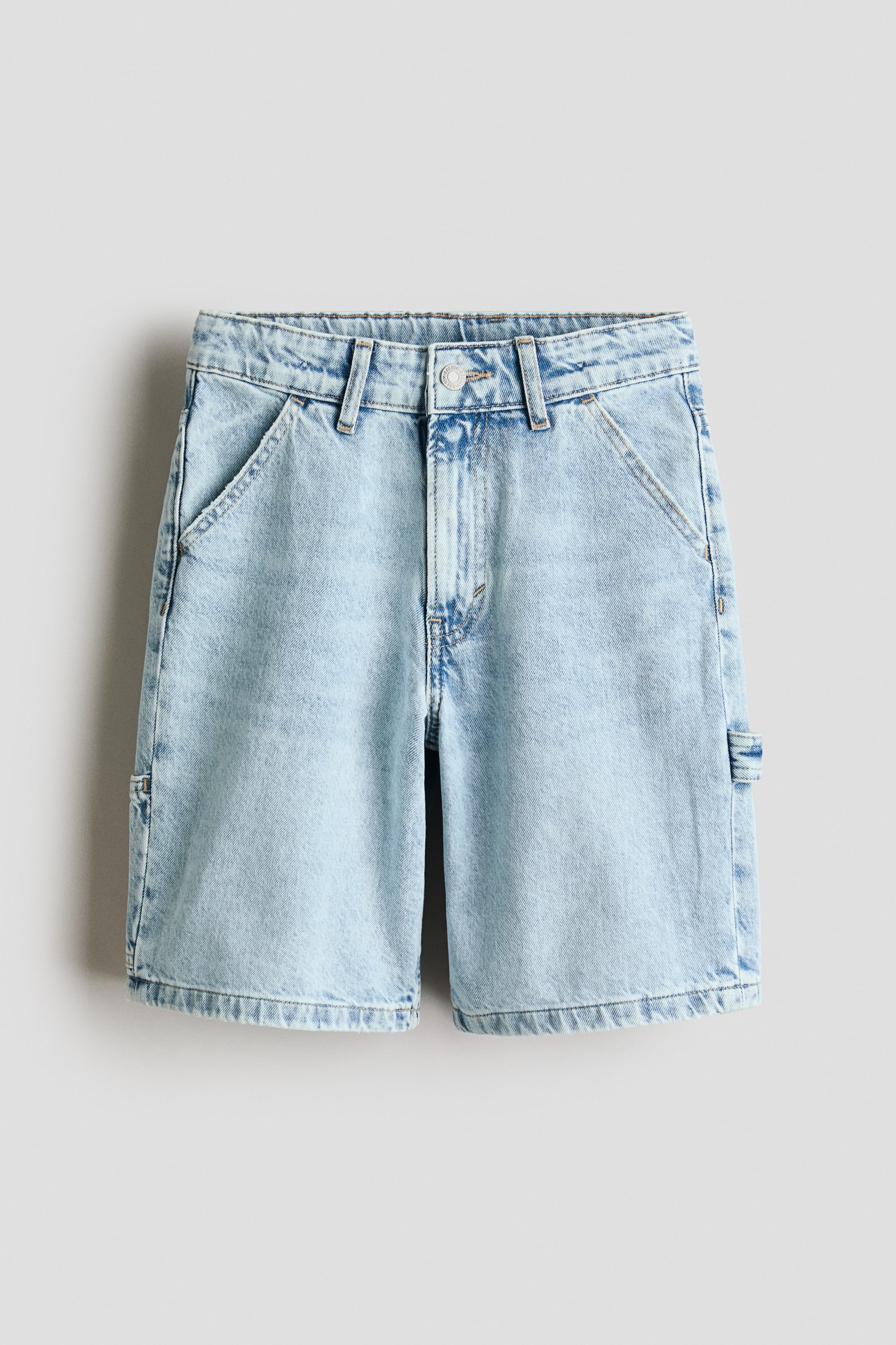 Agrandir l'image: Les shorts longueur genou sont présentés de face, en denim de coton bleu clair avec une coupe ample et une taille élastiquée ajustable. Ils sont conçus avec une braguette zippée et une fermeture à bouton, des poches avant en biais, et une boucle pour marteau sur le côté droit.