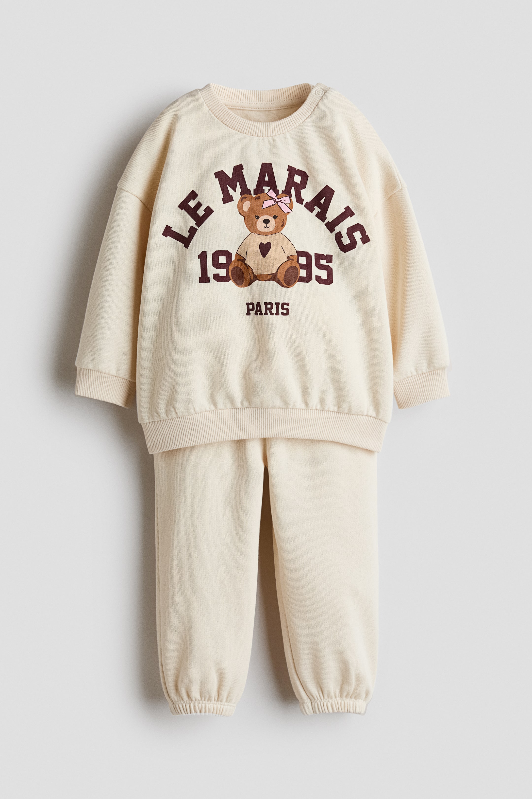 Nagyobb kép megtekintése: A krémszínű, kétszámos loungewear szett egy bordázott kerek nyakú, bordázott mandzsettájú és bordázott alsó szegélyű pulóvert tartalmaz, a bal vállán apró patentgombokkal. Az elejét egy rózsaszín masnival és szívvel díszített barna mackó grafika ékesíti, amelyet a „LE MARAIS 1995 PARIS” felirat vesz körül, míg a hozzá illő jogger nadrág gumírozott szárvégekkel teszi teljessé a szettet.