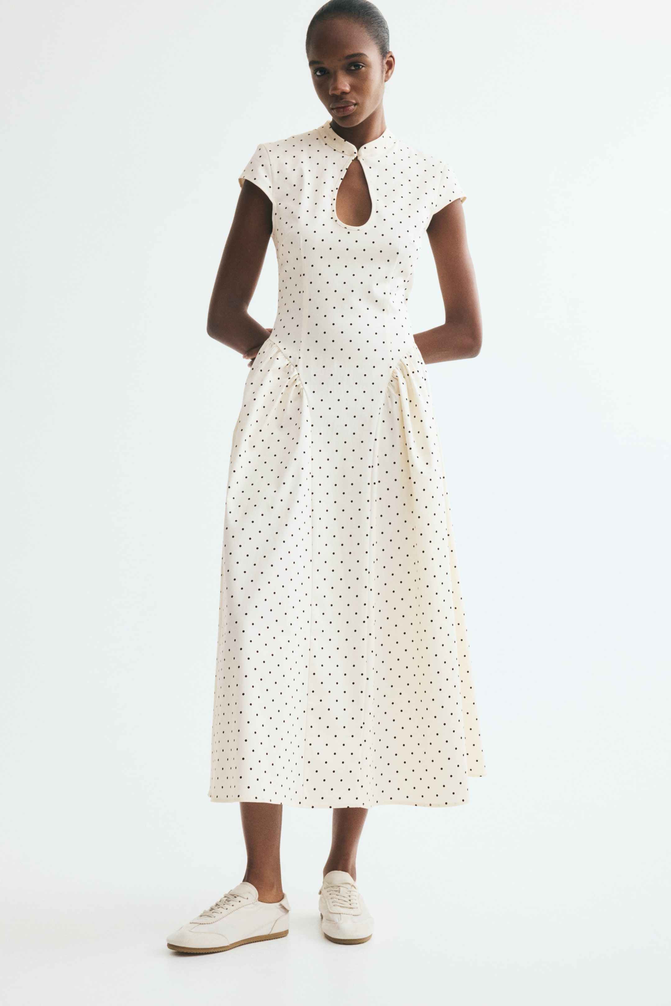 View larger image: A calf-length dress featuring a light background with black polka dots is being worn by the model. The dress has a band collar with a keyhole opening and button, cap sleeves, a fitted bodice, and a flared skirt with gathered inset panels at the hips. The model is also wearing light-coloured sneakers.