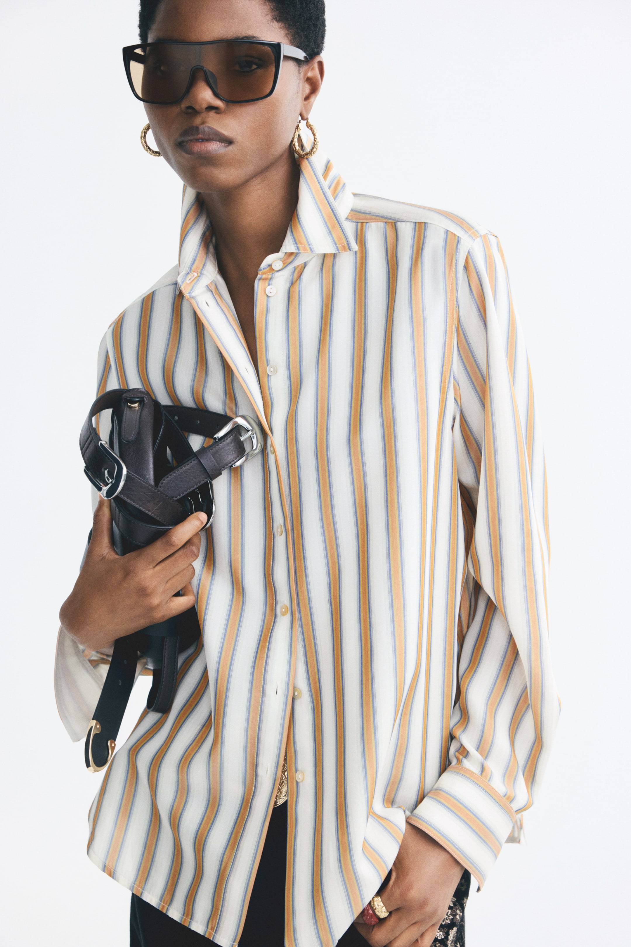 View larger image: A striped button-down shirt featuring vertical yellow, blue and white stripes, a pointed collar and long sleeves is being worn by the model. The shirt is styled partially unbuttoned at the collar, and the model is holding a black leather bag with silver buckles.