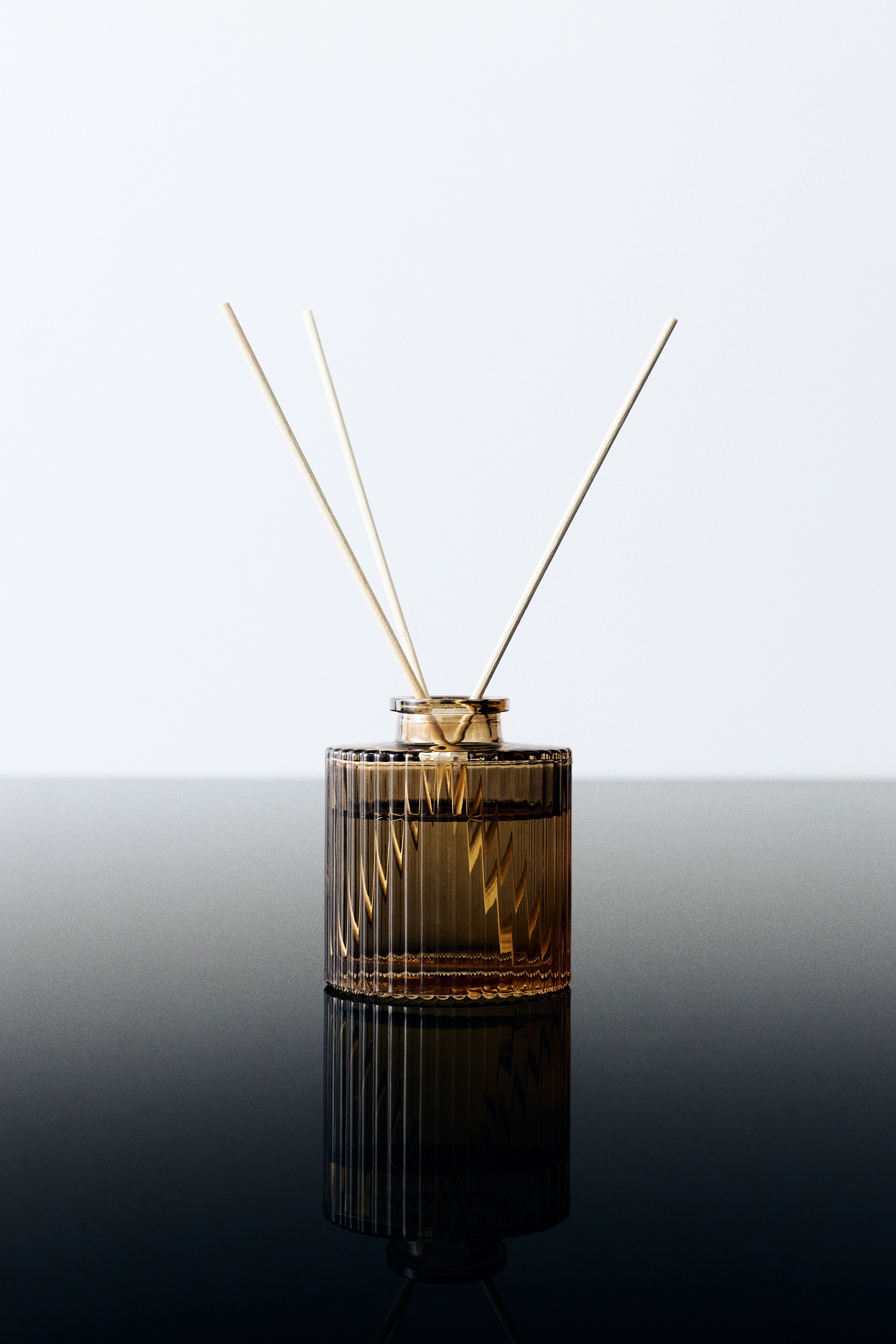 Un diffuseur à roseaux est montré de face, avec une bouteille cylindrique en verre côtelé teintée dâambre et remplie de liquide. Cinq tiges en bois de couleur claire se dressent dans la bouteille, disposées en éventail à partir dâun collet de couleur dorée au niveau du goulot.