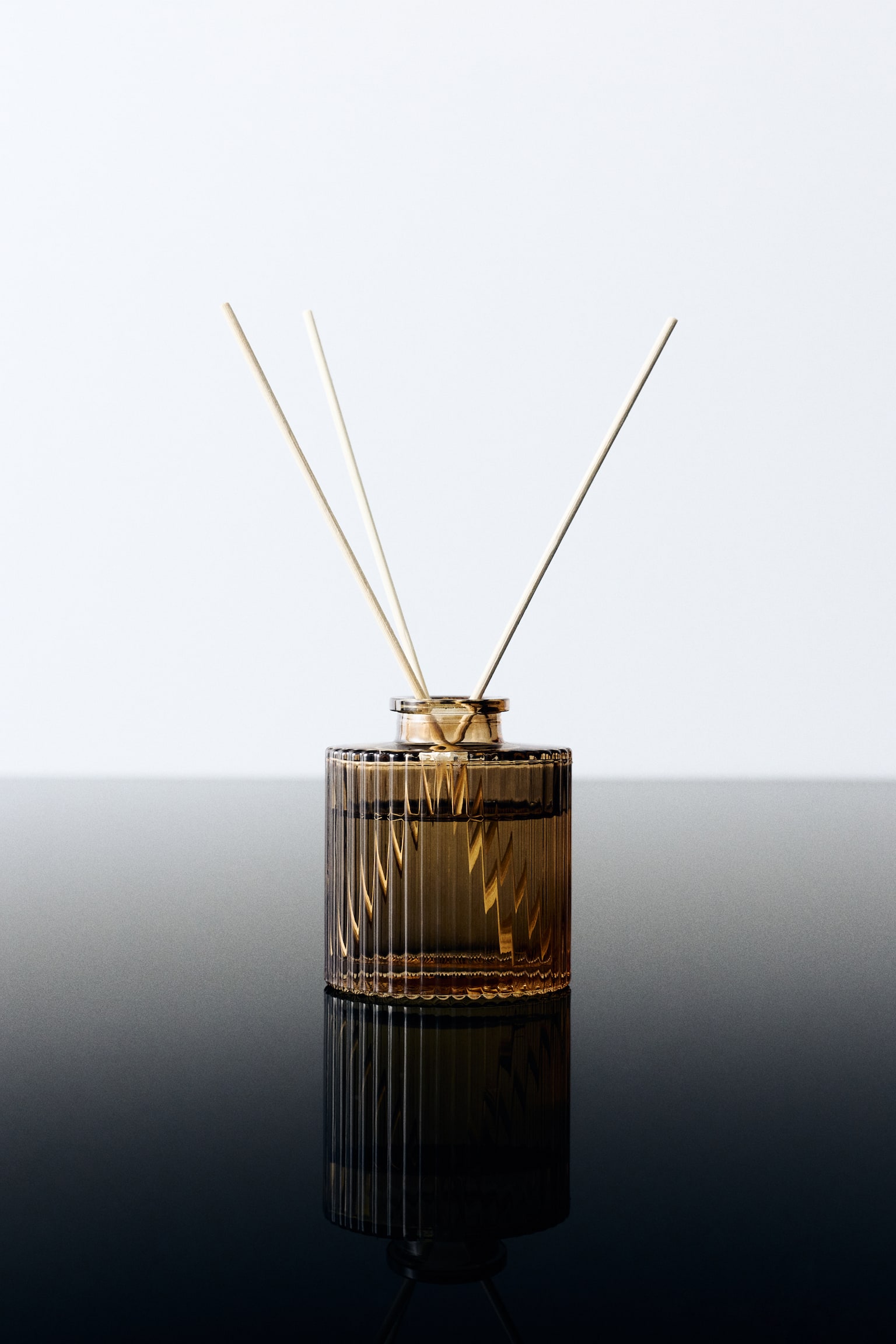 Un diffuseur à bâtonnets mettant en scène un flacon cylindrique en verre cannelé teinté ambré, rempli de liquide, présenté de face. Trois tiges en bois clair reposent dans l’ouverture étroite du flacon et s’évasent vers le haut. - 1