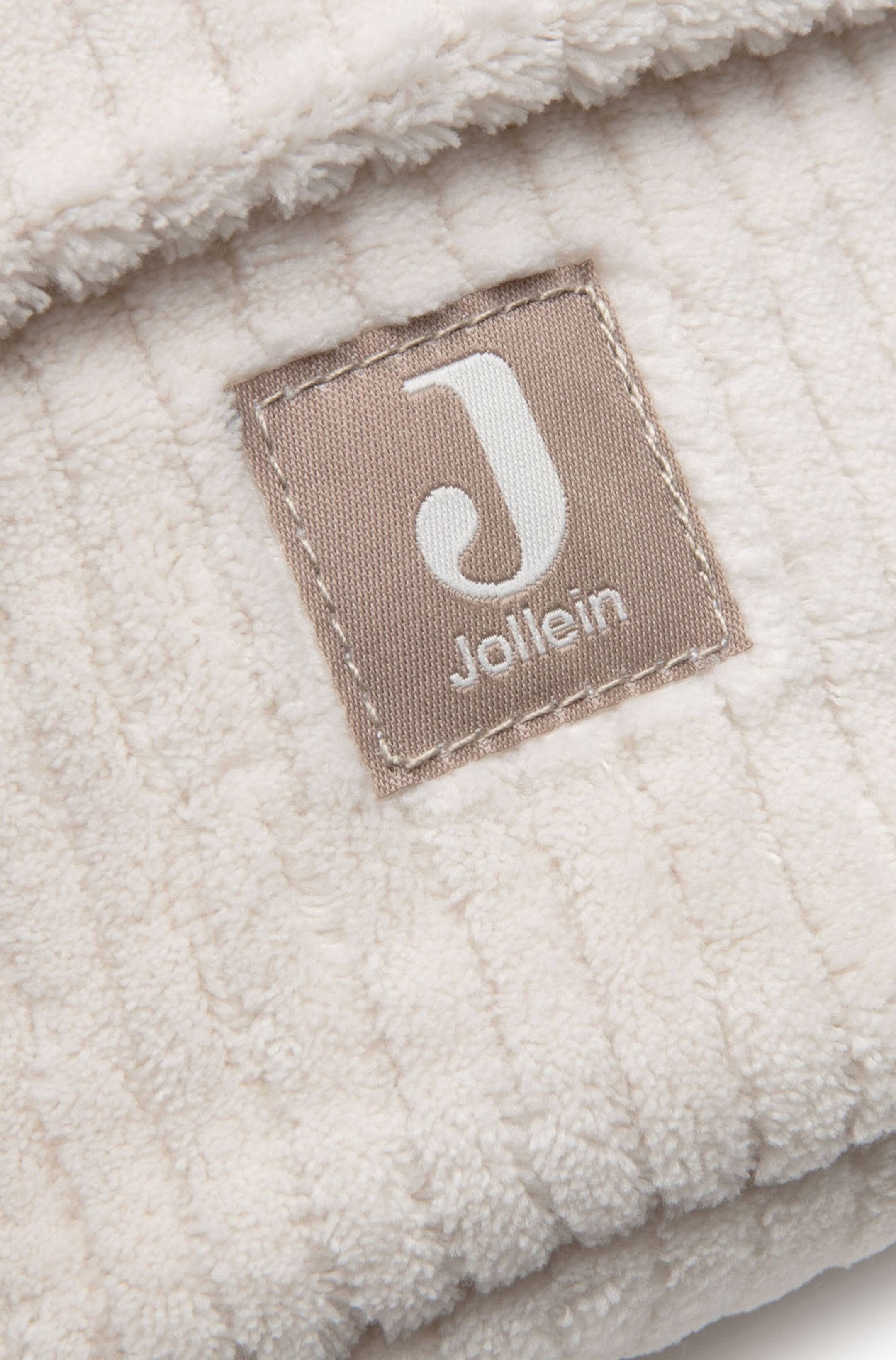 Agrandir l'image: Un produit textile en peluche de couleur crème présente une texture côtelée et un léger pli dans son matériau. Une étiquette en tissu rectangulaire beige, arborant l'inscription "Jollein" et un 'J' stylisé en broderie blanche, est cousue sur le produit.