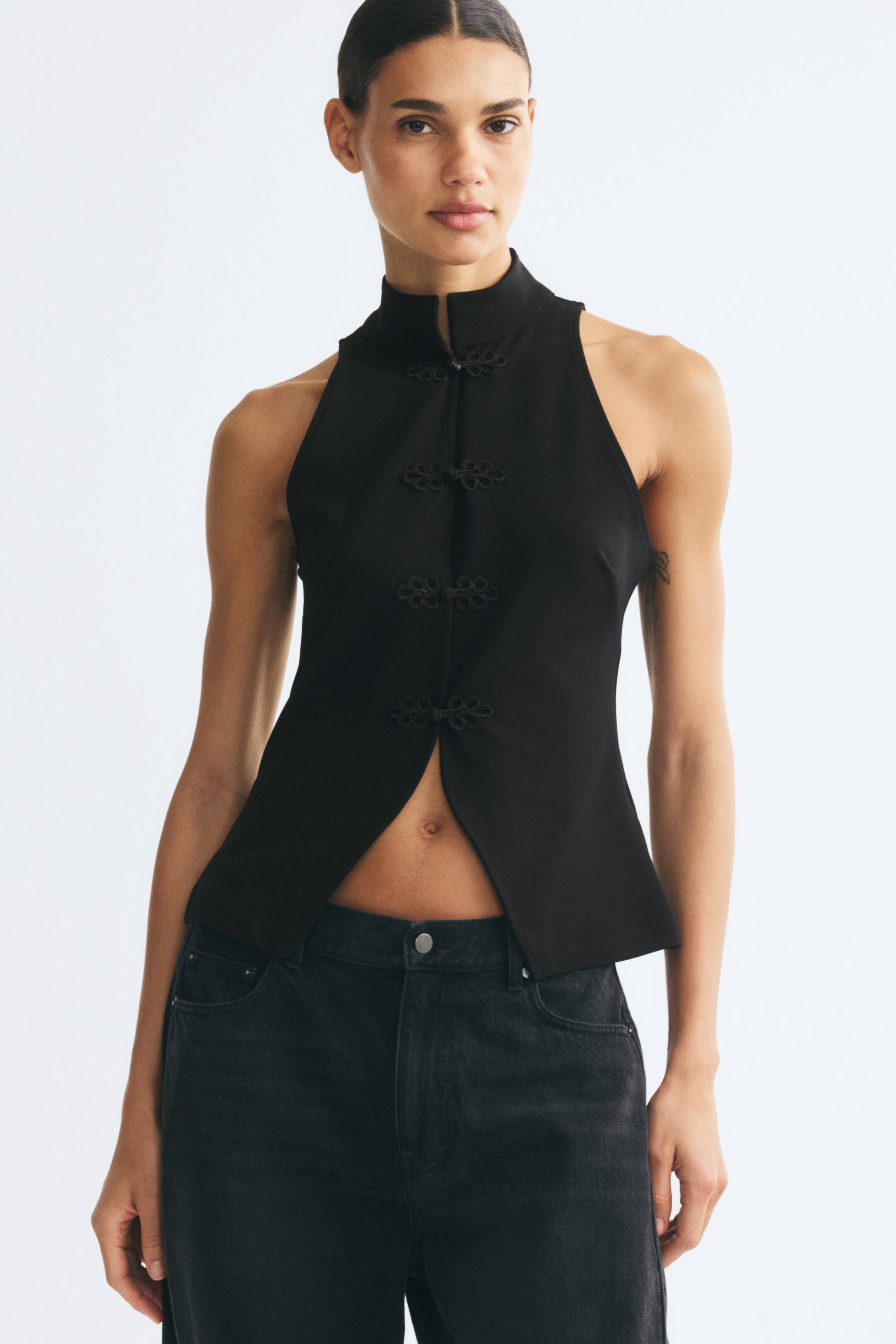 Visualizza immagine più grande: La modella indossa un gilet nero senza maniche in jersey interlock pesante, caratterizzato da un colletto alto alla coreana e da quattro bottoni decorativi con nodo di corda che chiudono il davanti. Il gilet è abbinato a jeans in denim nero, mettendo in risalto un orlo spaccato che si apre leggermente in vita.
