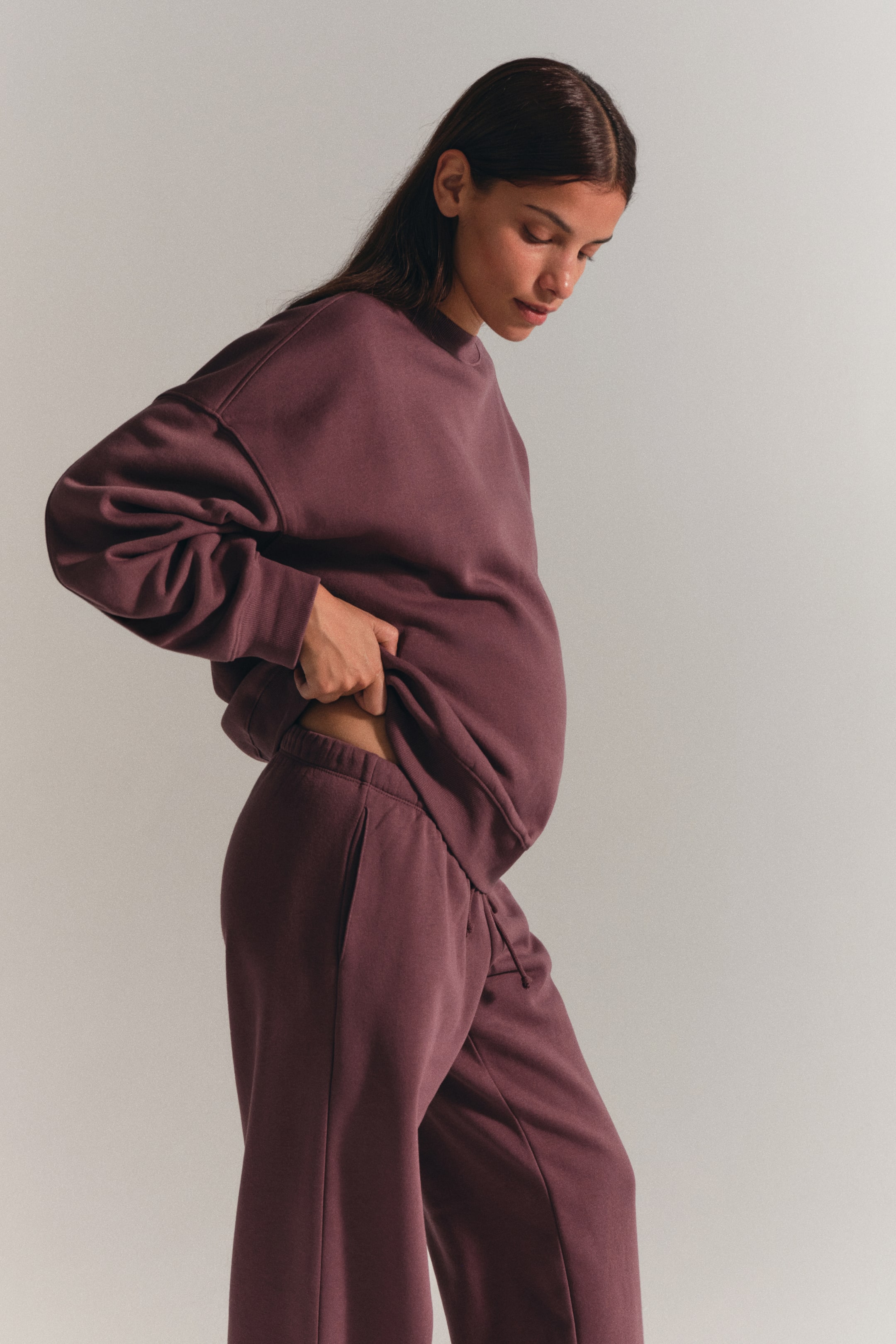 View larger image: A matching dark plum sweatsuit is worn by the model, featuring a crewneck sweater with dropped shoulders and ribbed cuffs, paired with matching pants with an elastic waistband. The model is lifting the hem of the sweater with one hand to adjust the fit and reveal the waistband of the pants.