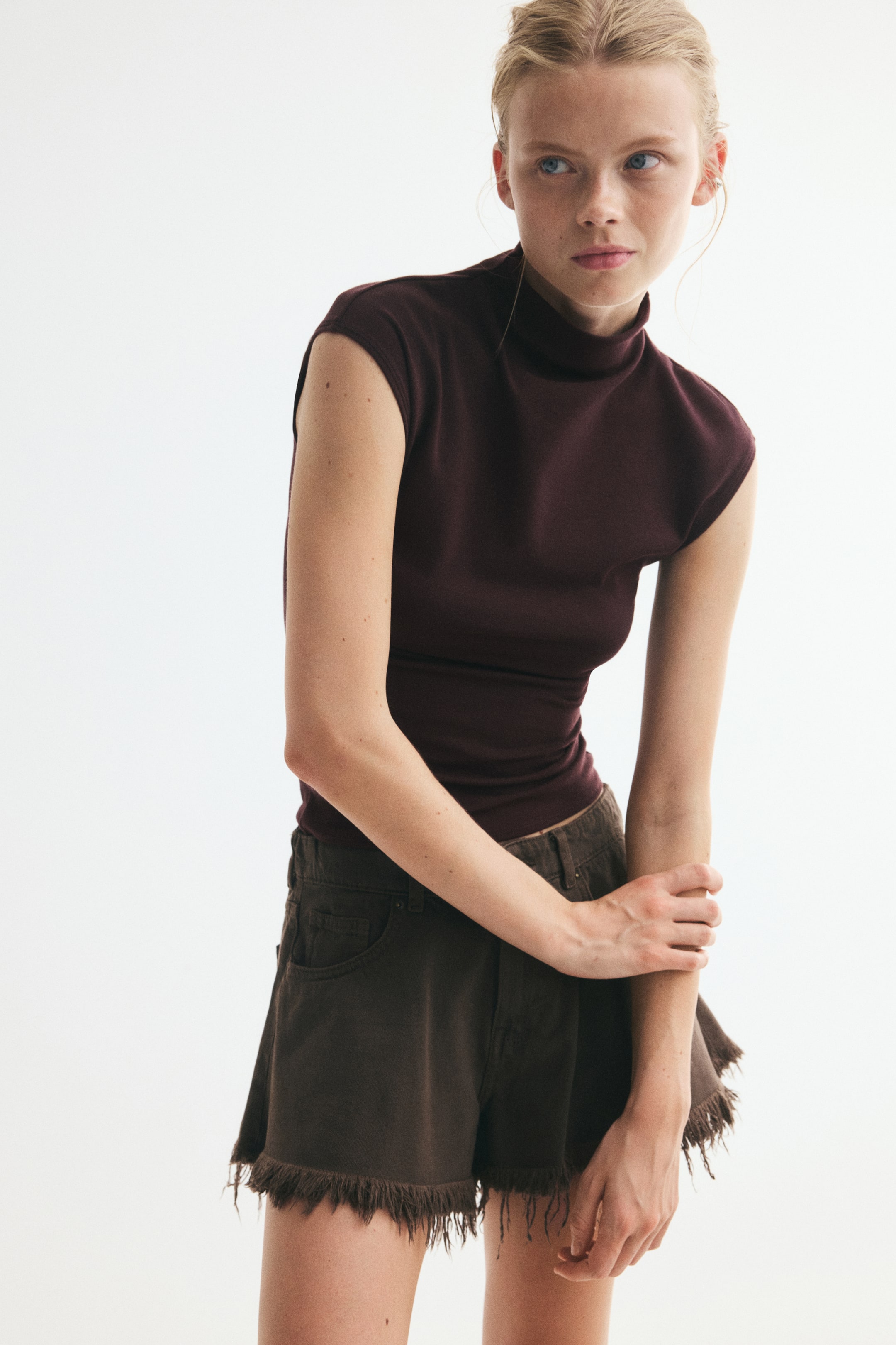 View larger image: The model is wearing a deep burgundy slim-fit top with a stand collar and cap sleeves. The top is styled with dark brown denim shorts featuring frayed hems, and the model is holding one arm with her hand.