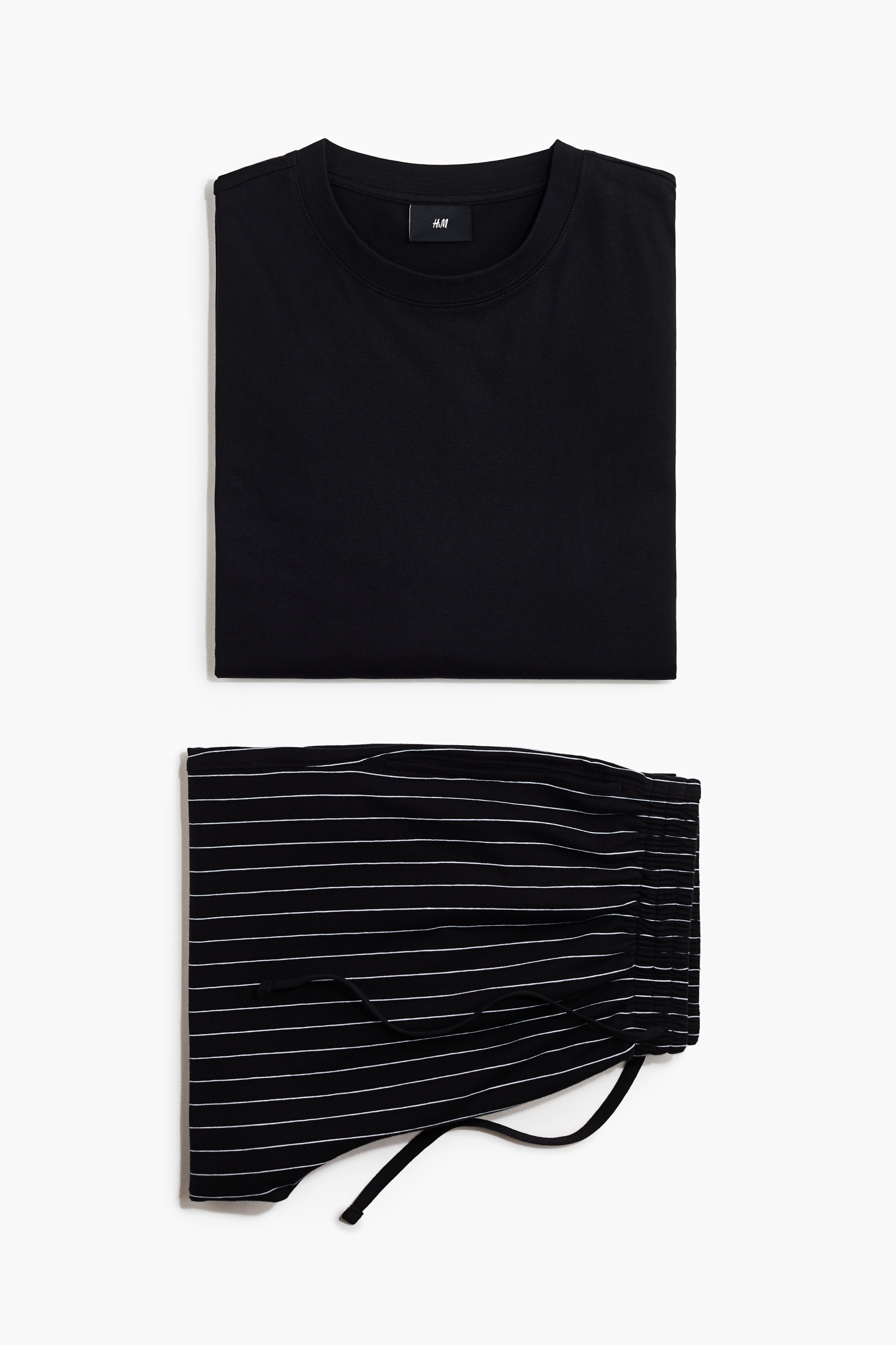 Un ensemble de pyjama composÃ© dâun t-shirt noir uni Ã  coupe dÃ©contractÃ©e, avec un col rond, et dâun pantalon noir assorti Ã  coupe droite, ornÃ© de fines rayures blanches verticales, est portÃ© par le mannequin. Le mannequin est debout, pieds nus, les mains dans les poches latÃ©rales du pantalon.