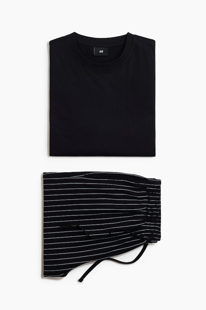 Een opgevouwen pyjamaset ligt plat, met een effen zwart T-shirt met ronde hals boven een paar opgevouwen zwarte broeken. De broeken hebben dunne, horizontale witte strepen en een elastische tailleband met een zwart trekkoord. - 1