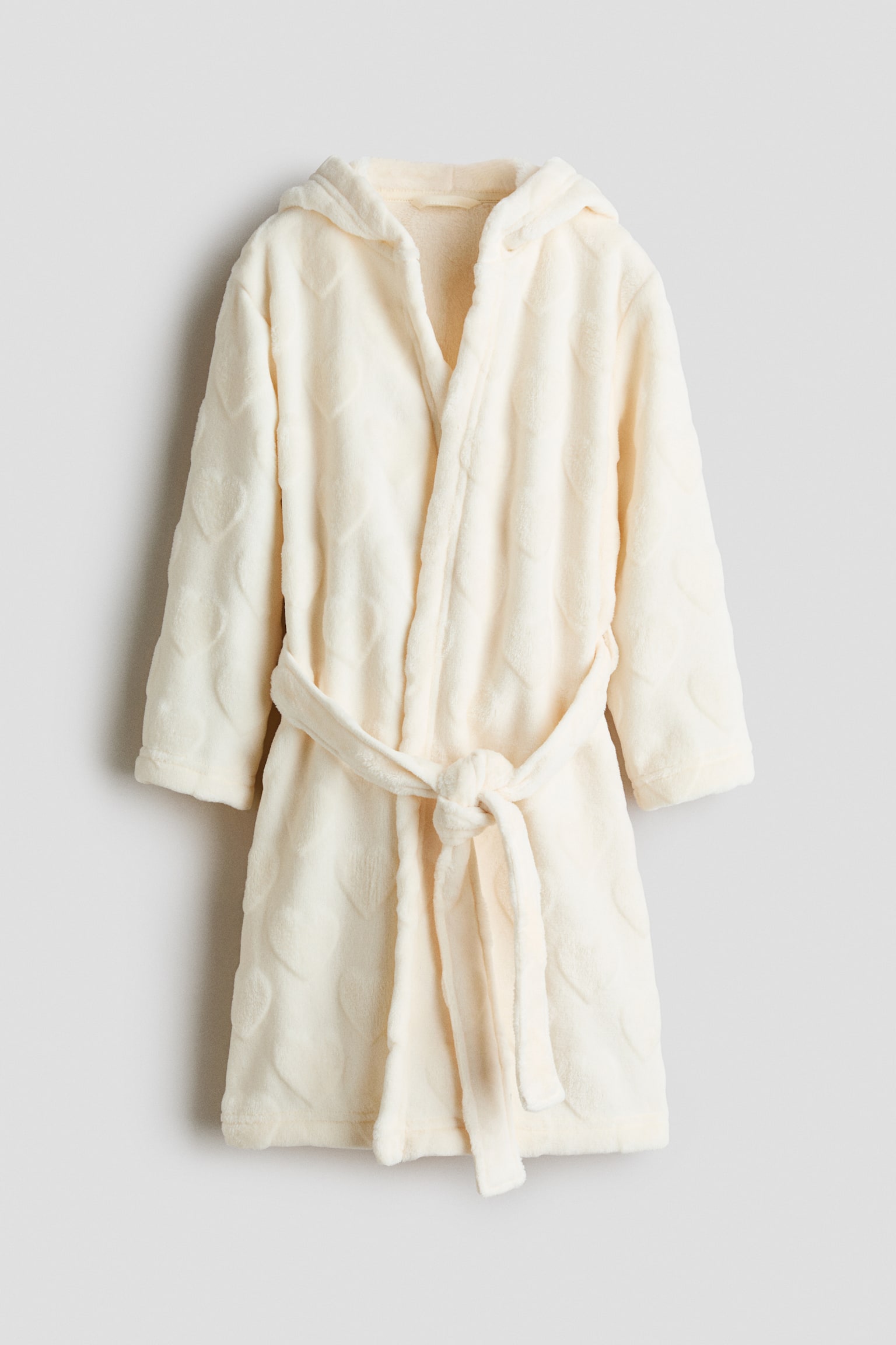 Een vooraanzicht van een crÃ¨mekleurige badjas van zachte fleece hangt vlak, met een all-over reliÃ«f hartpatroon, een capuchon en een bijpassende ceintuur die in de taille is geknoopt.