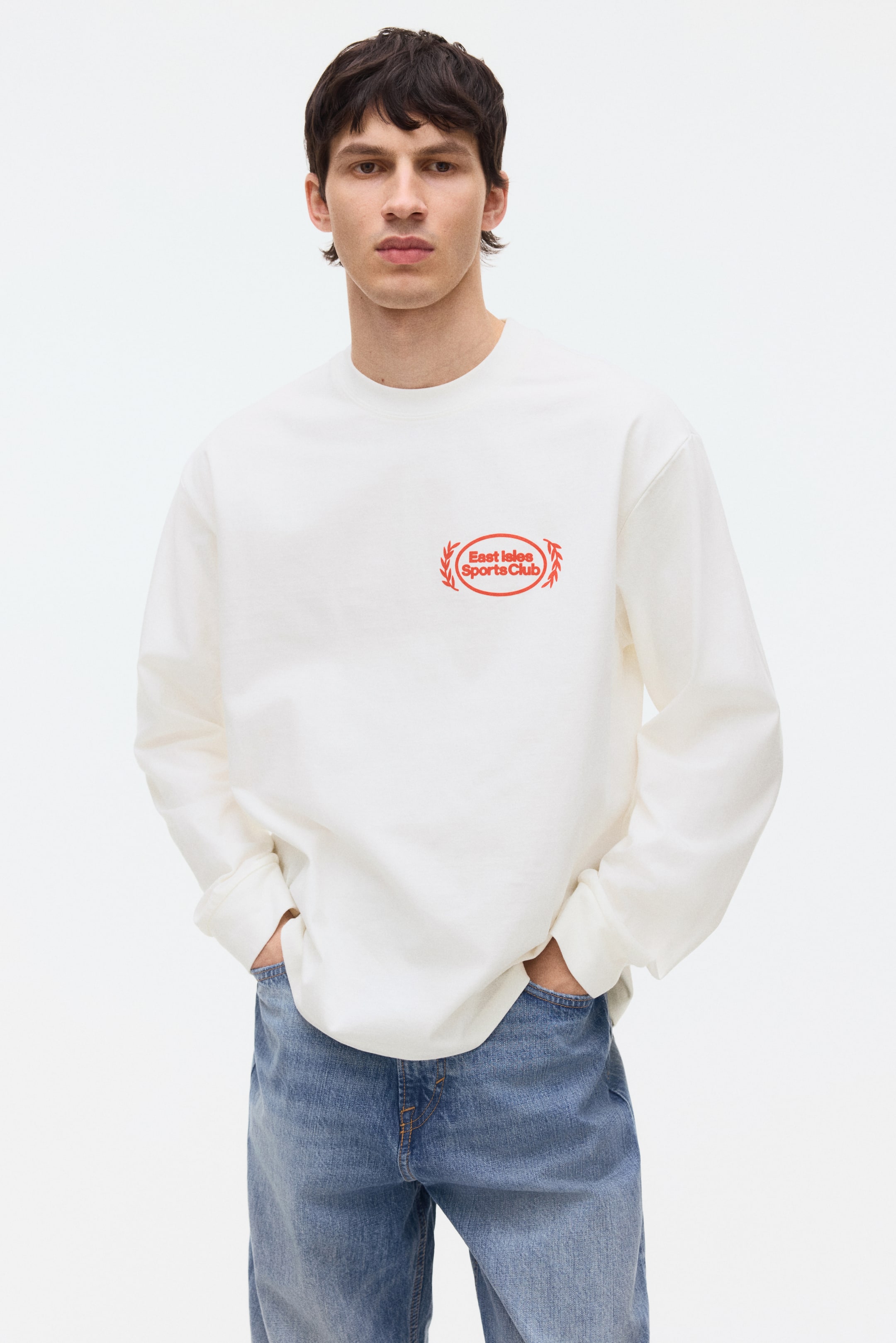 הצגת תמונה גדולה יותר: סווטשירט לבן עם צווארון עגול מציג לוגו אובלי אדום "East Isles Sports Club" על החזה השמאלי שלו. הוא מעוצב עם שרוולים ארוכים, חפתים מצולעים ושוליים מצולעים, ומציג גזרה רפויה.
