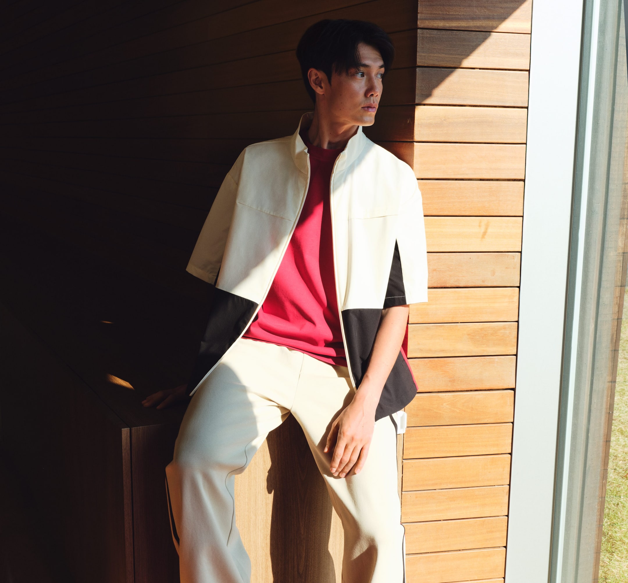 View larger image: A cream short-sleeved jacket with a stand collar and front zipper is worn open over a red crew-neck shirt. The jacket features black side panels, and cream pants with a dark side stripe complete the ensemble.