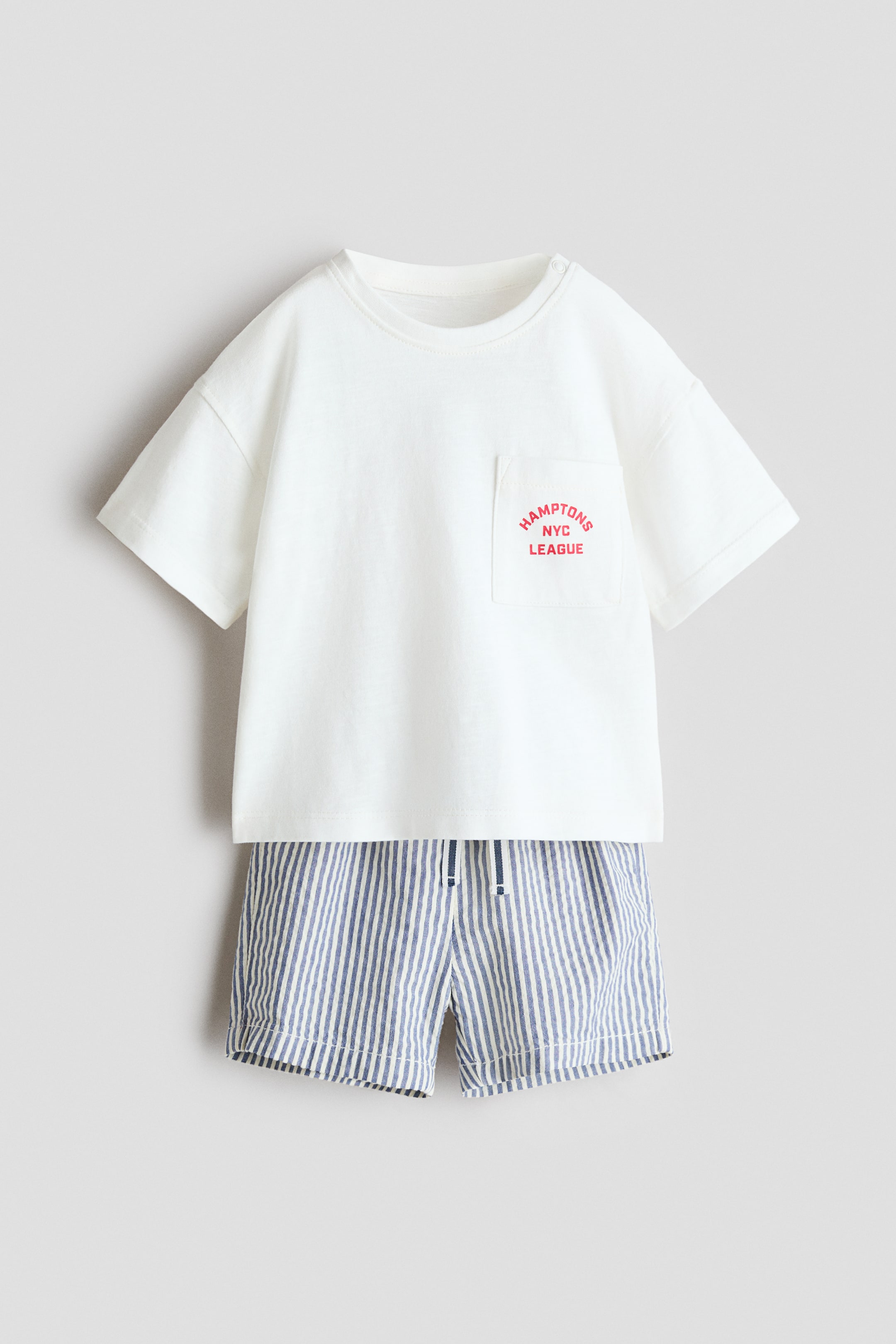 View larger image: A two-piece outfit showcasing a white, short-sleeved tee and blue-and-white striped shorts, both front-facing. The tee highlights a ribbed crew neck and a left chest pocket adorned with red 'HAMPTONS NYC LEAGUE' text, while the shorts feature a blue drawstring at the waist.