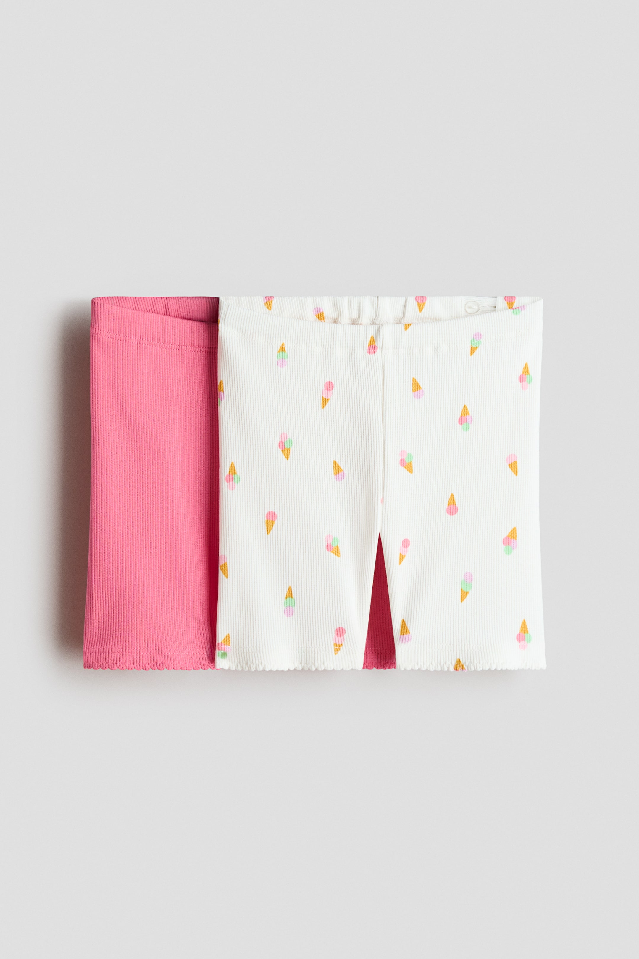 Agrandir l'image: Deux paires de shorts de cyclisme sont présentées en vue de face. Une paire est d'un rose uni, tandis que l'autre est blanche avec un motif coloré de cônes de glace. Les deux paires sont confectionnées en jersey de coton doux et côtelé, avec une taille élastiquée ajustable et des bords festonnés aux ourlets.