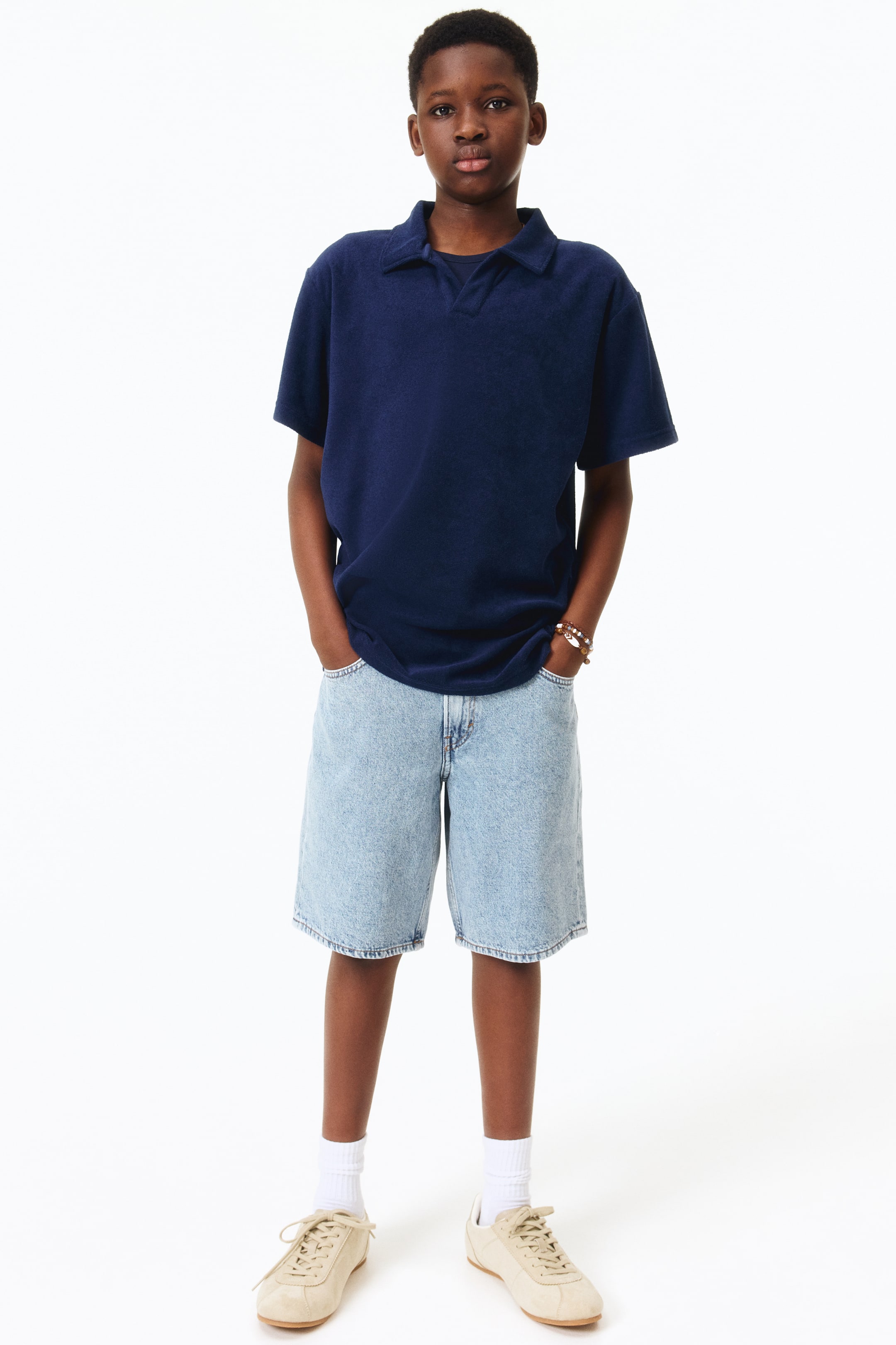 Se større billede: En marineblå kortærmet poloshirt med en spredt krave og V-hals placket bæres, med en blød, tekstureret strik. Den er stylet med lyseblå løstsiddende denimshorts, hvide crew-sokker og beige snøresneakers, alle præsenteret fra en frontvisning.