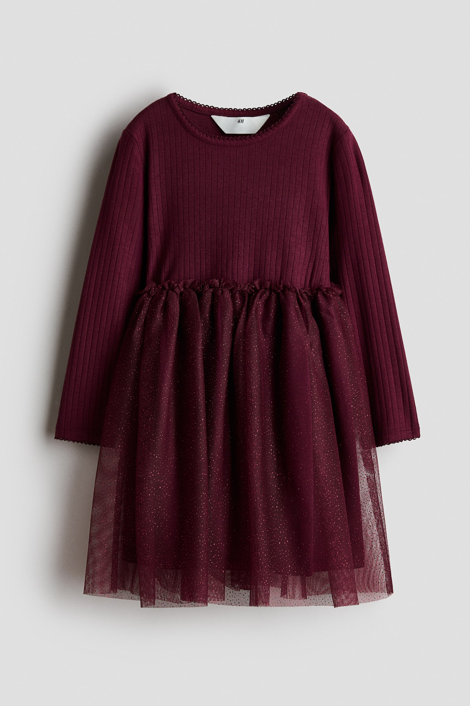 A dark burgundy dress is shown facing front, featuring a long-sleeved, ribbed jersey bodice with a round neckline and picot trims at the collar and cuffs. The dress is designed with a small frill trim at the waist and a flared, lined tulle skirt that sparkles with fine glitter.
