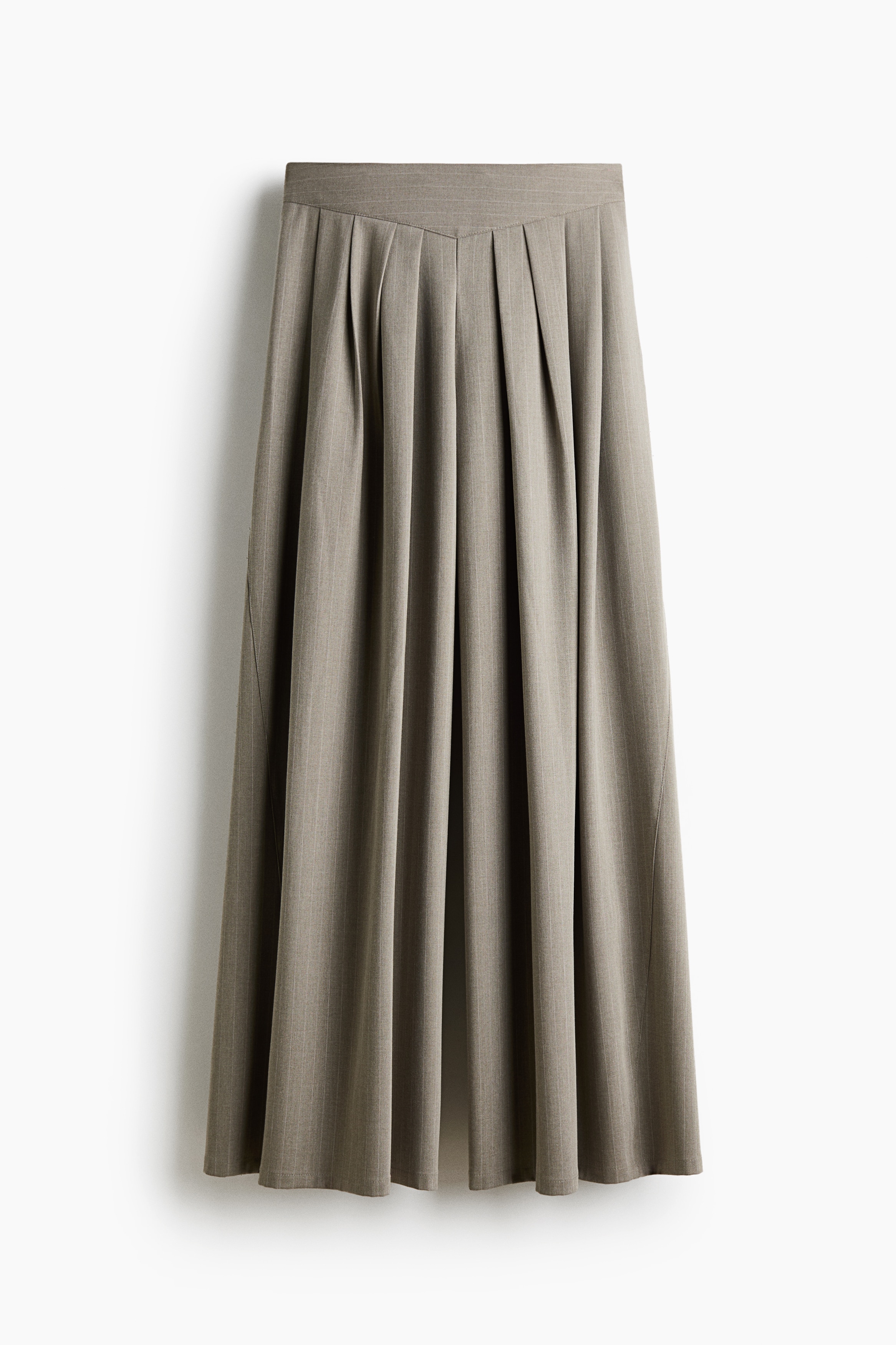 Loose-fit twill pants in a light taupe shade are worn by the model, featuring a high waist, prominent front pleats, and a wide-leg silhouette. The pants are styled with a layered white shirt under a brown knit sweater and paired with white pointed-toe shoes.