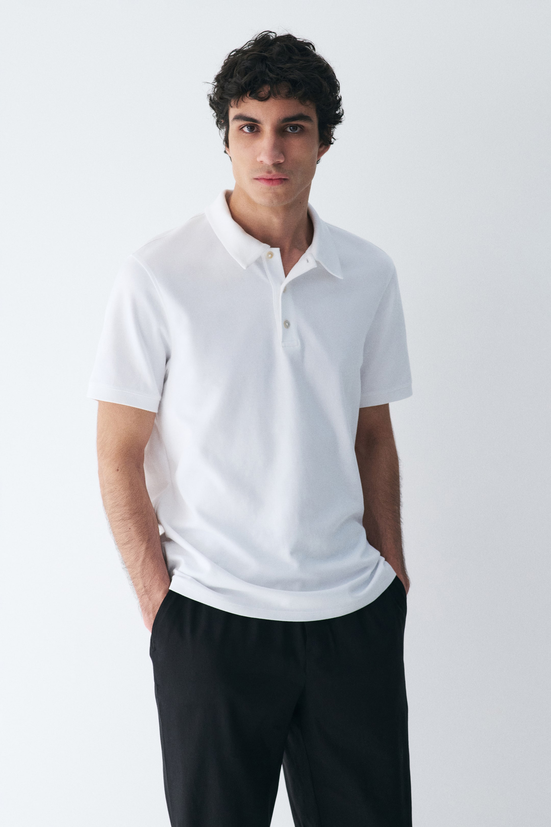 View larger image: A white polo shirt is showcasing a classic spread collar and a two-button placket with light-coloured buttons. It features short, hemmed sleeves and fits loosely on the body, facing forward.