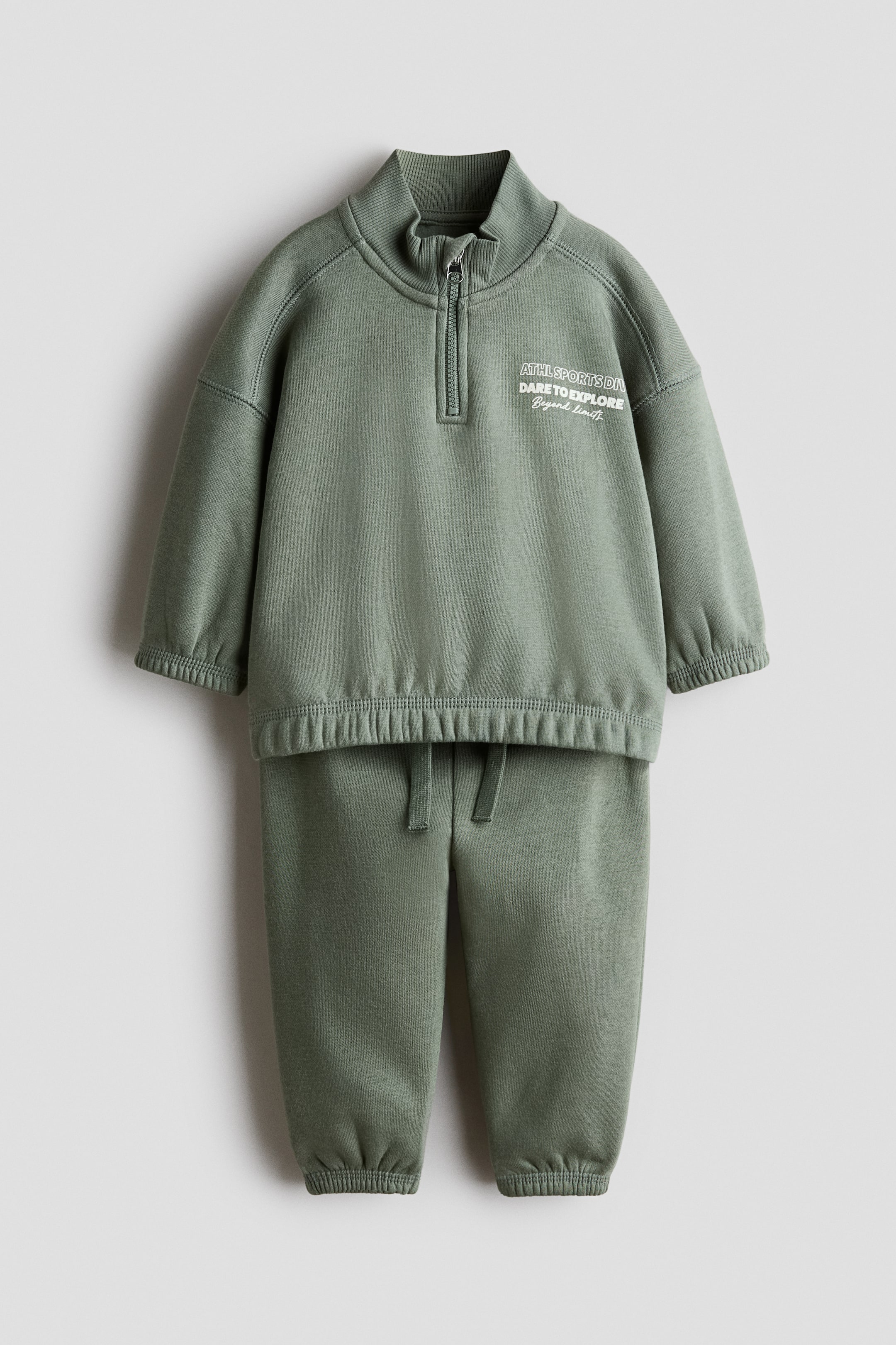 Zobacz większy obraz: Zielony dwuczęściowy dres jest prezentowany i składa się z bluzy z półzamkiem oraz dopasowanych spodni joggerów. Bluza ma prążkowany stójkowy kołnierz, zapięcie na ćwierćzamek, elastyczne mankiety, elastyczny dół oraz biały nadruk tekstowy „ATHL SPORTS DIV DARE TO EXPLORE Beyond limits” na klatce piersiowej, natomiast spodnie joggery mają pas ze sznurkiem do regulacji i elastyczne ściągacze przy kostkach.